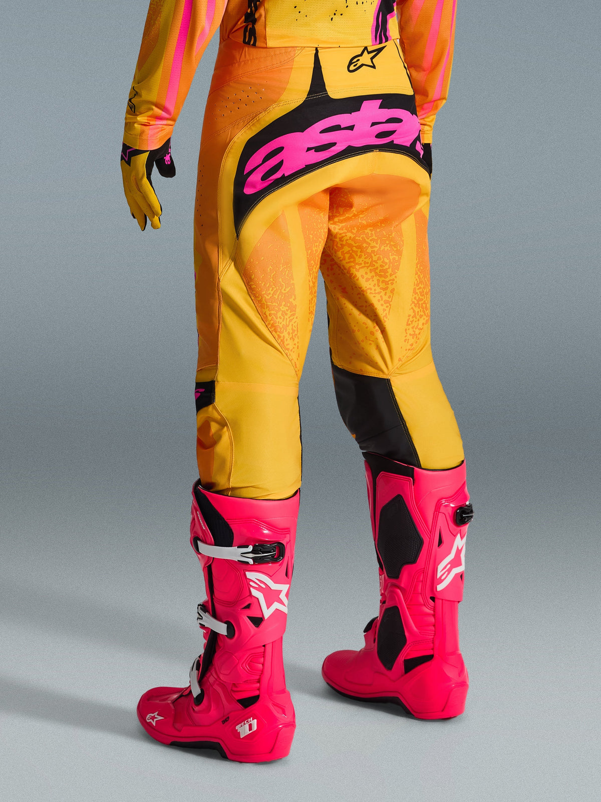 Spodnie Alpinestars 2026 Techstar Nomur, spodnie motocrossowe, pomarańczowe, żółte i różowe, widok z tyłu, z dużym różowym logotypem „astars” na siedzeniu, w połączeniu z jaskraworóżowymi butami Tech 10 i pasującą koszulką, profesjonalna odzież wyścigowa do motocrossu
