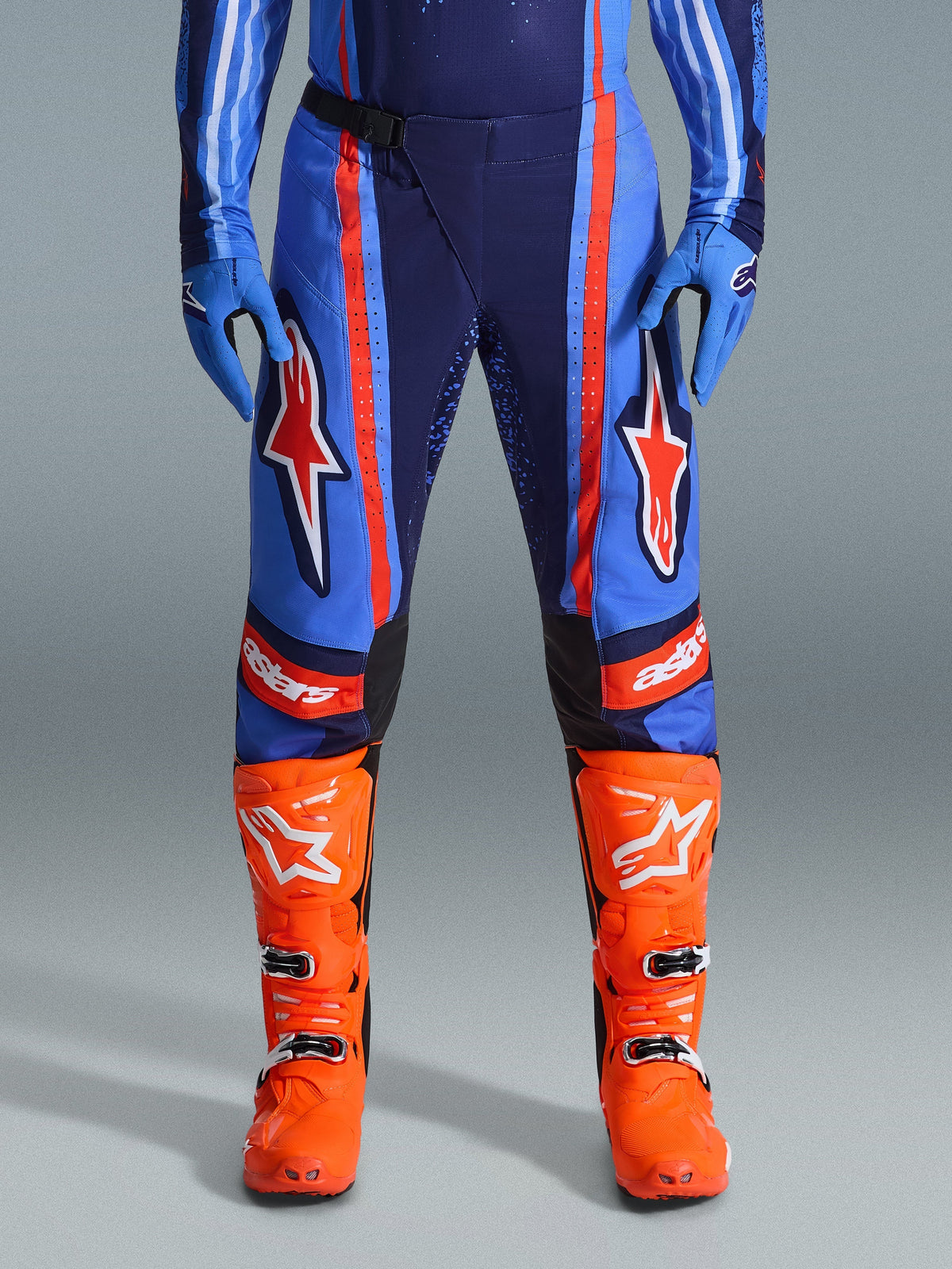 Alpinestars 2026 Techstar Nomur Pants, spodnie motocrossowe, ciemnogranatowe, pomarańczowe i błękitne UCLA, z wyrazistymi pomarańczowymi pasami i dużym białym logo gwiazdy na udach, noszone z jaskrawopomarańczowymi butami wyścigowymi i niebieskimi rękawicami, sportowy krój wyścigowy