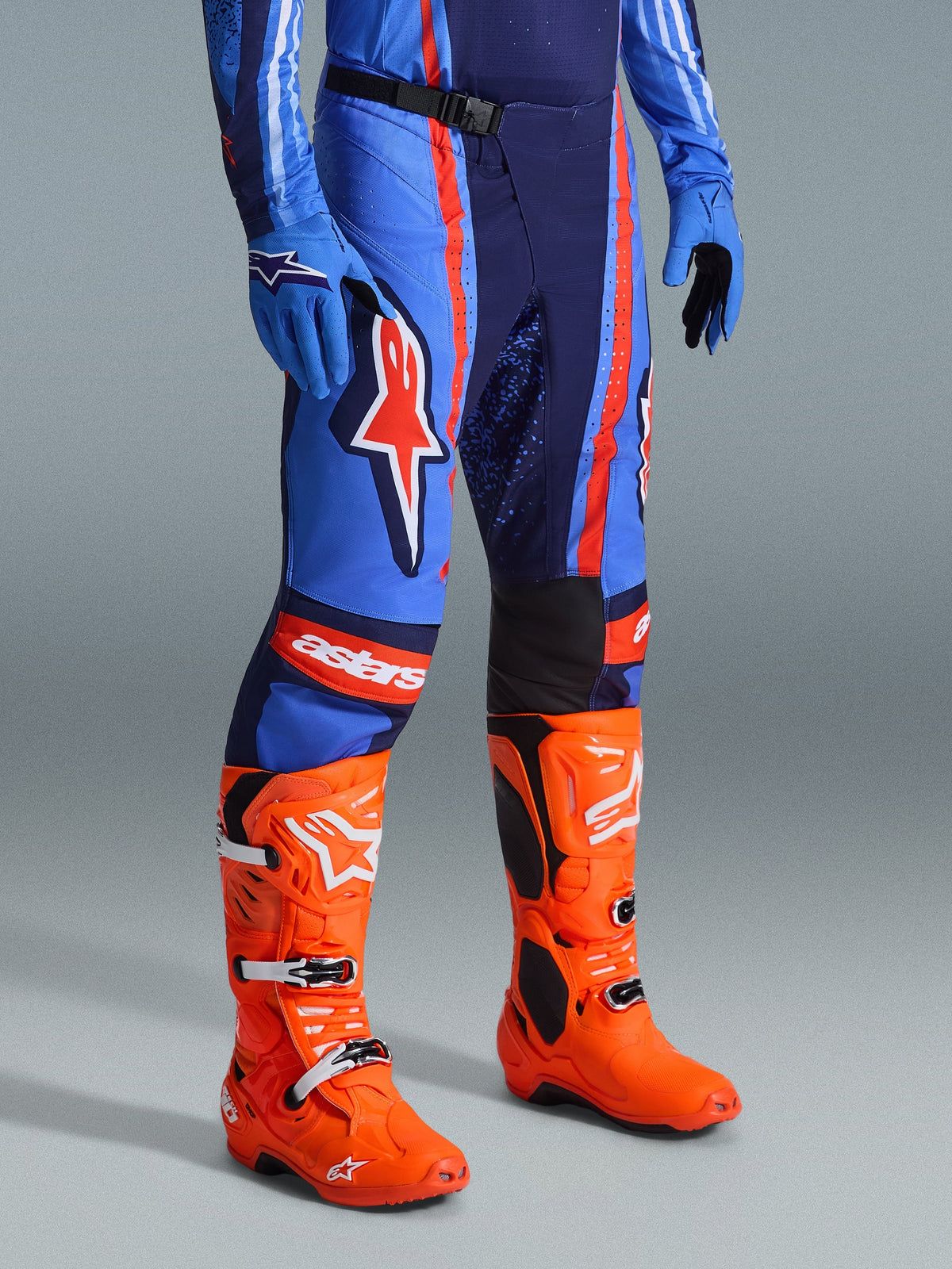 Alpinestars 2026 Techstar Nomur Pants, spodnie motocrossowe, granatowe, pomarańczowe i niebieskie UCLA, noszone z jaskrawopomarańczowymi butami motocrossowymi i pasującym niebieskim strojem, wyrazisty branding, wielomateriałowa konstrukcja zapewniająca wydajność w terenie