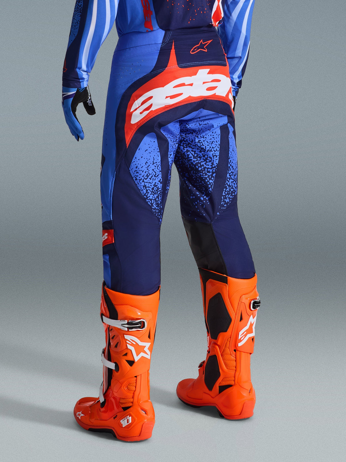 Alpinestars 2026 Techstar Nomur Pants, spodnie motocrossowe, ciemnogranatowe, pomarańczowe i błękitne (UCLA Blue), widok z tyłu z białym logo „Astars” na siedzeniu, noszone z jaskrawopomarańczowymi butami motocrossowymi i pasującą koszulką, sportowy krój wyścigowy do rywalizacji off-road.