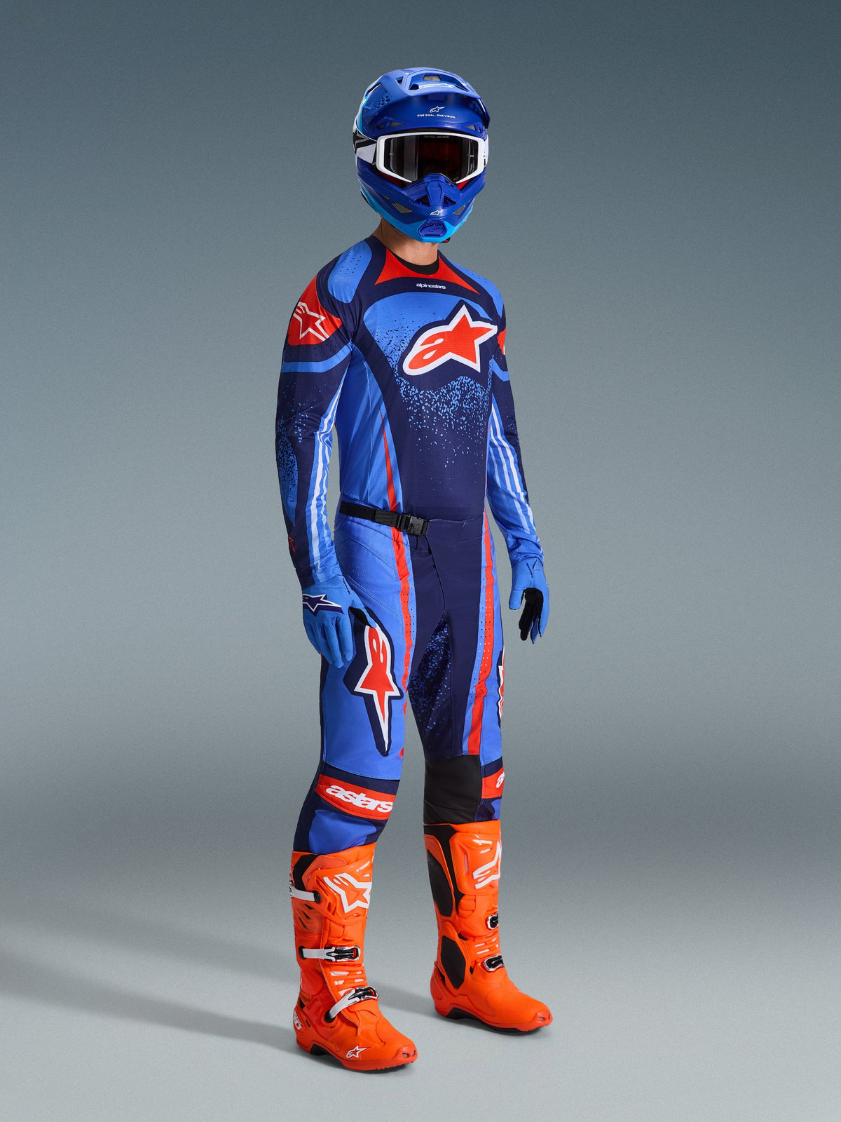 Alpinestars 2026 Techstar Nomur Pants, spodnie motocrossowe, ciemnogranatowe, pomarańczowe i błękitne (UCLA Blue), noszone przez zawodnika w pełnym stroju wyścigowym, w tym pasująca koszulka, niebieski kask i jaskrawopomarańczowe buty, zaprojektowane do profesjonalnych wyścigów terenowych