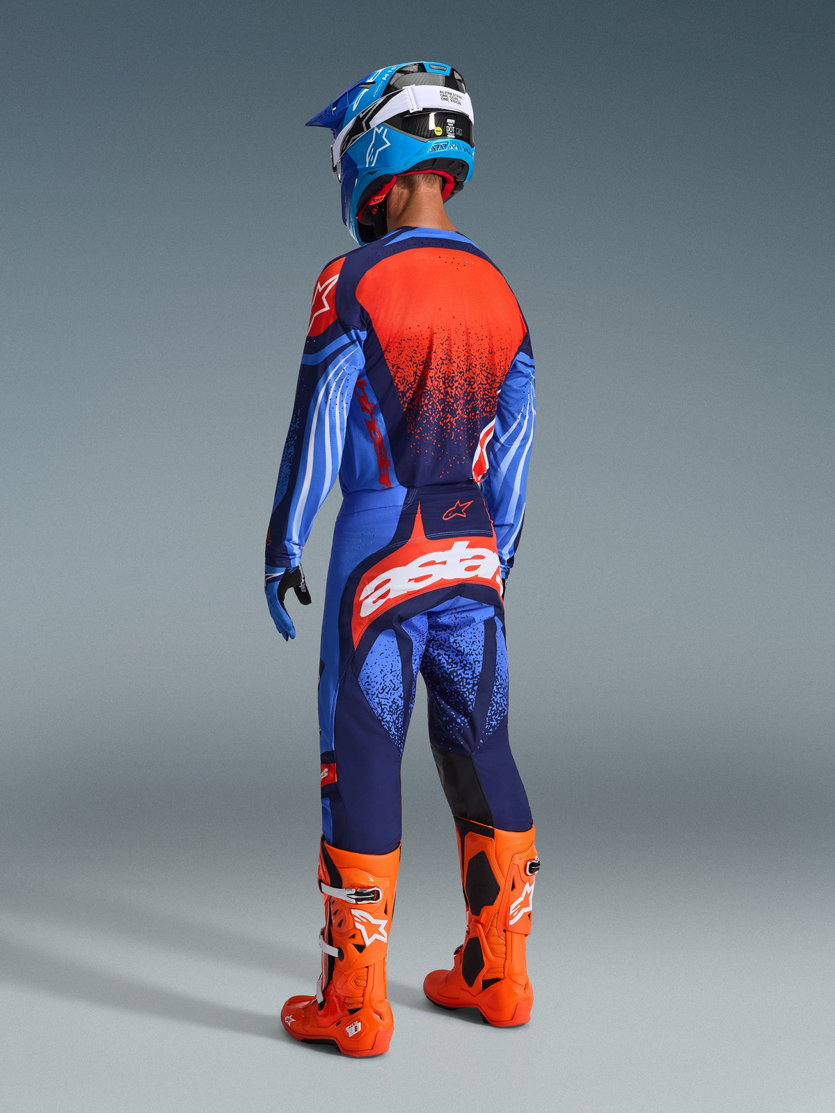 Alpinestars Techstar Nomur Pants 2026, spodnie motocrossowe, ciemnogranatowe, pomarańczowe i błękitne (UCLA Blue), widok od tyłu zawodnika w pełnym stroju, duże białe logo na siedzeniu, jaskrawopomarańczowe buty, niebieska koszulka i kask, profesjonalny strój motocrossowy.
