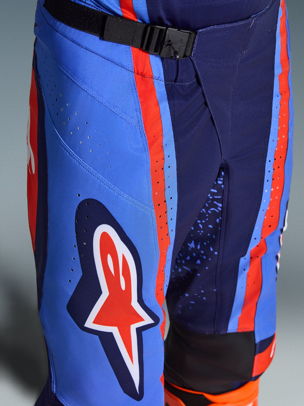 Alpinestars Techstar Nomur Pants 2026, spodnie motocrossowe w kolorach ciemnogranatowym, pomarańczowym i błękitnym, zbliżenie na magnetyczne zapięcie SX2 w pasie, laserowo wycinane perforacje na udzie oraz kultowe logo gwiazdy, wytrzymała odzież off-road