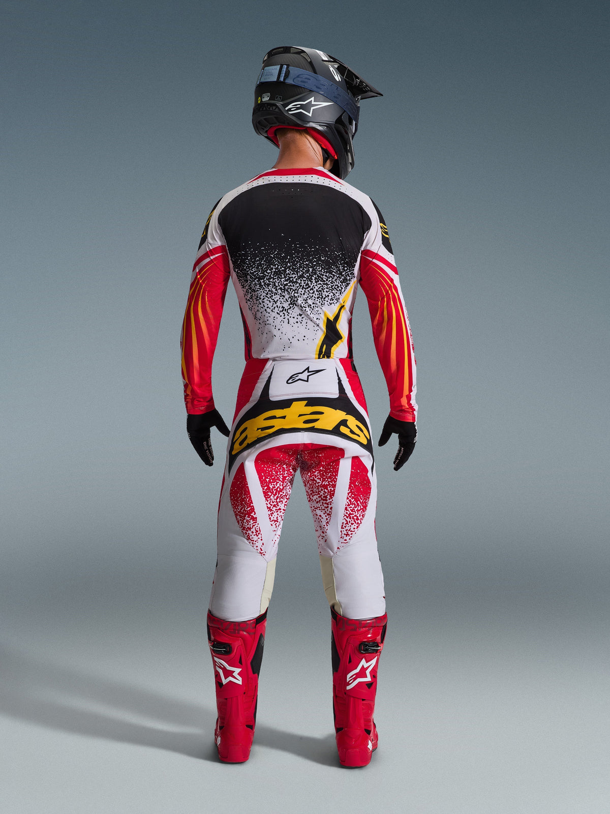 Widok z tyłu zawodnika motocrossowego ubranego w koszulkę Alpinestars 2026 Techstar Nomur w kolorach czarnym, czerwonym i białym z żółtymi akcentami, połączoną z dopasowanymi spodniami motocrossowymi z wyrazistym żółtym brandingiem, jaskrawoczerwonymi butami wyścigowymi i czarnym kaskiem, tworzącymi kompletny zestaw odzieży do wyścigów off-road.