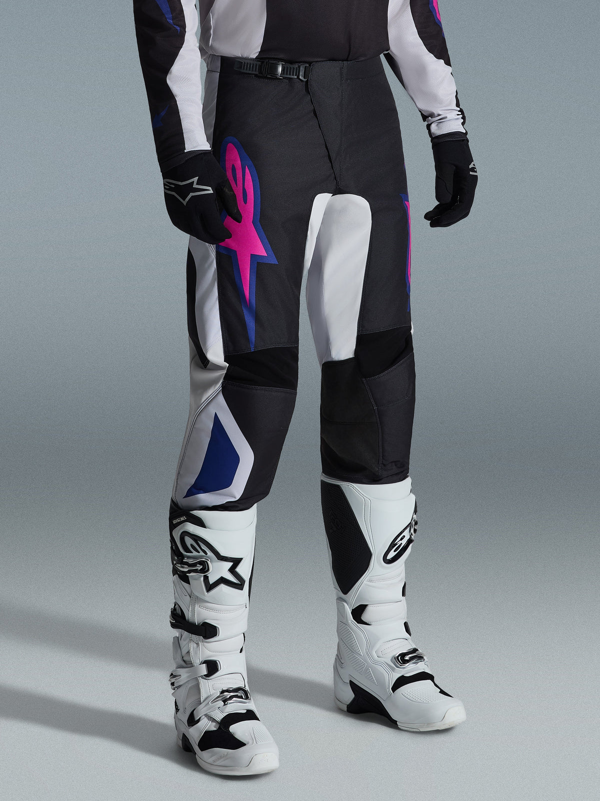 Alpinestars Fluid Grid Pants 2026, spodnie motocrossowe, czarne, jasnoszare i fioletowe, z wyrazistym różowym logo gwiazdy i niebieskimi geometrycznymi akcentami, noszone z białymi butami motocrossowymi i czarnymi rękawicami, ergonomiczna odzież do jazdy terenowej