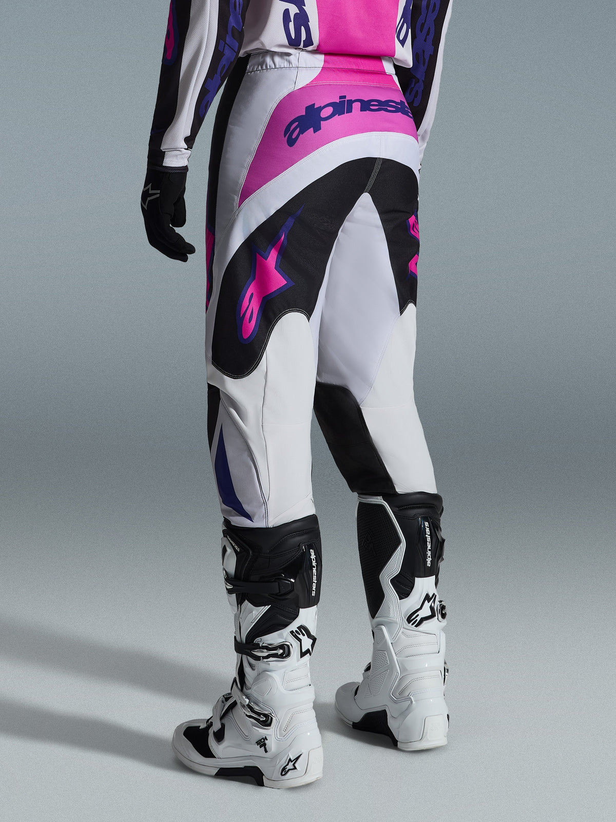 Spodnie Alpinestars 2026 Fluid Grid, spodnie motocrossowe, czarne, jasnoszare i fioletowe, widok od tyłu motocyklisty w spodniach i biało-czarnych butach, różowe i fioletowe logo Alpinestars, sportowy krój do wyścigów off-road