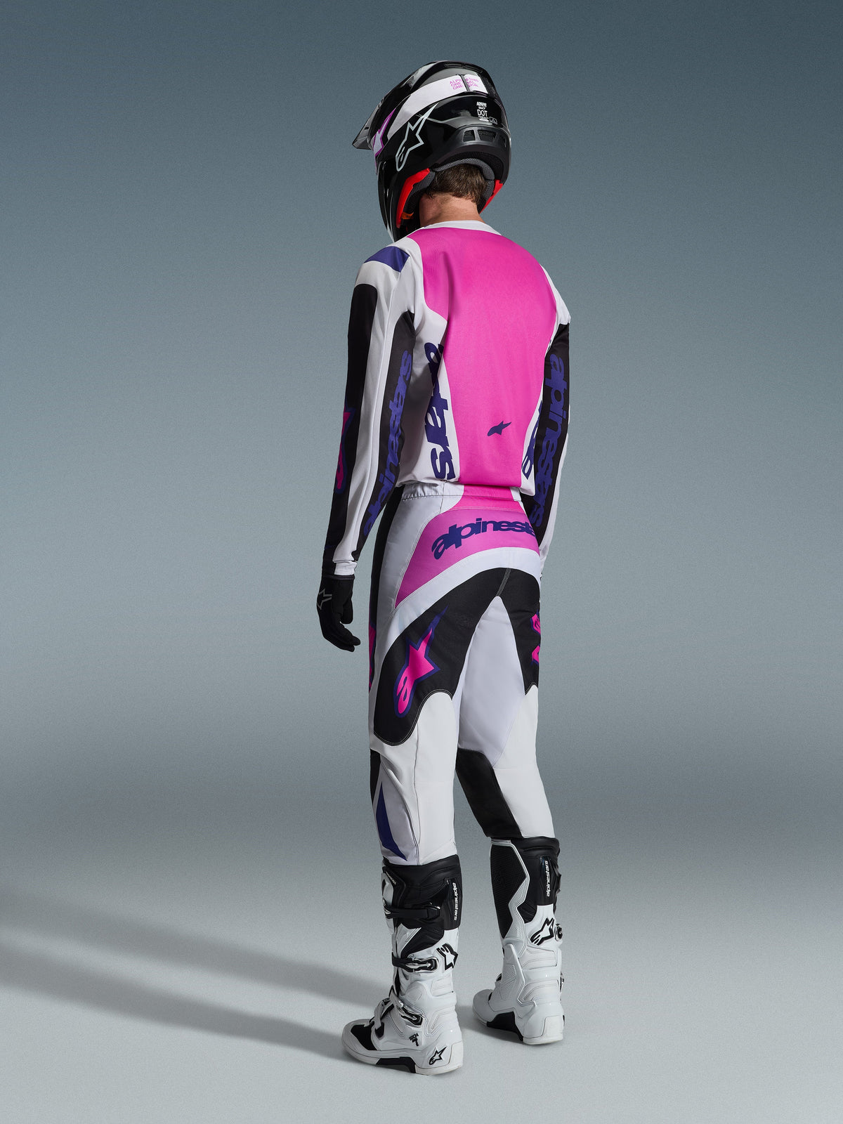 Spodnie Alpinestars Fluid Grid 2026, spodnie motocrossowe, czarne, jasnoszare i fioletowe, widok od tyłu na motocyklistę w pasującej koszulce i białych butach, z wyrazistymi fioletowymi logotypami i jaskrawymi różowymi panelami, zaprojektowane z myślą o wydajności w motocrossie terenowym.