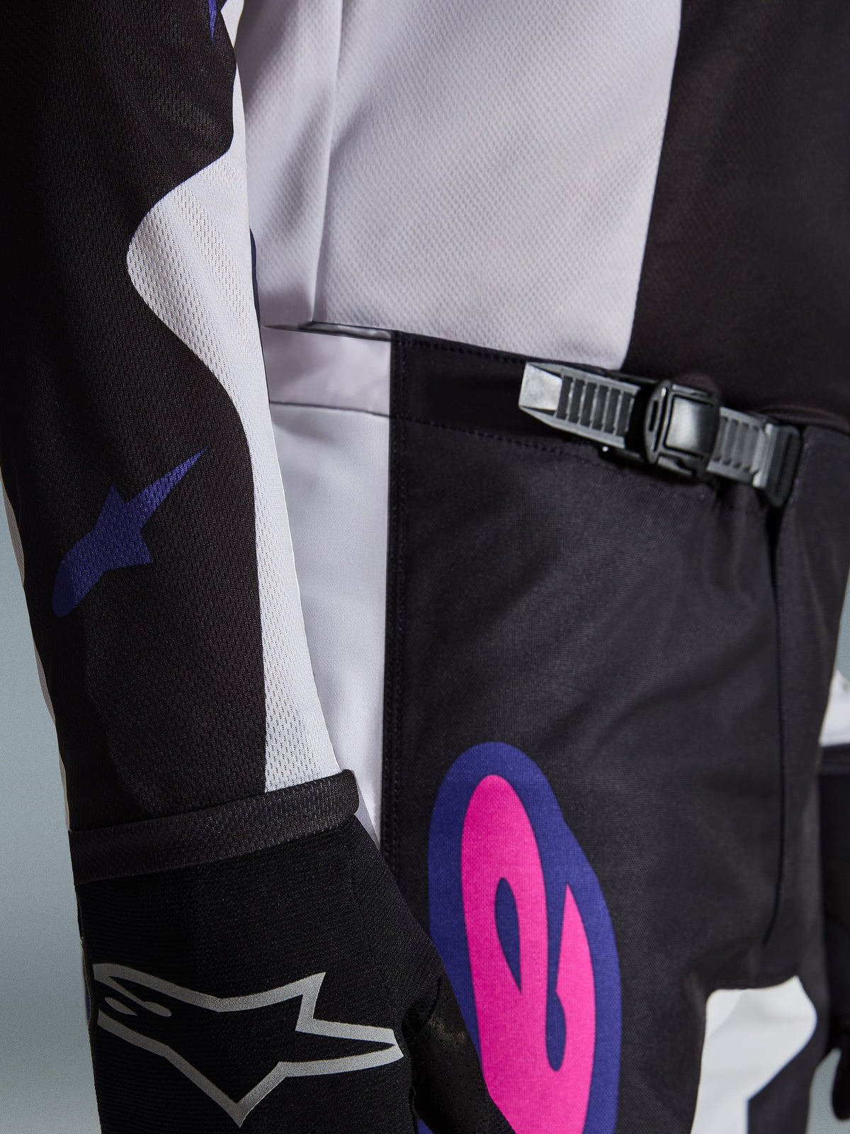 Alpinestars 2026 Fluid Grid Pants, spodnie motocrossowe, czarne, jasnoszare i fioletowe, zbliżenie na system zapięcia na zapadkę w pasie oraz wyrazistą różowo-fioletową grafikę na biodrze, noszone z pasującą bluzą i czarną rękawicą.