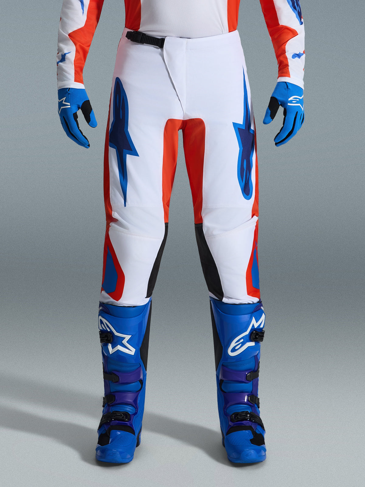 Spodnie Alpinestars Fluid Grid 2026, spodnie motocrossowe, pomarańczowe i niebieskie (UCLA Blue), zestaw z pasującymi niebieskimi butami i rękawicami, biała konstrukcja z wyrazistymi niebieskimi logo i pomarańczowymi akcentami, ergonomiczny design zapewniający wydajność w terenie