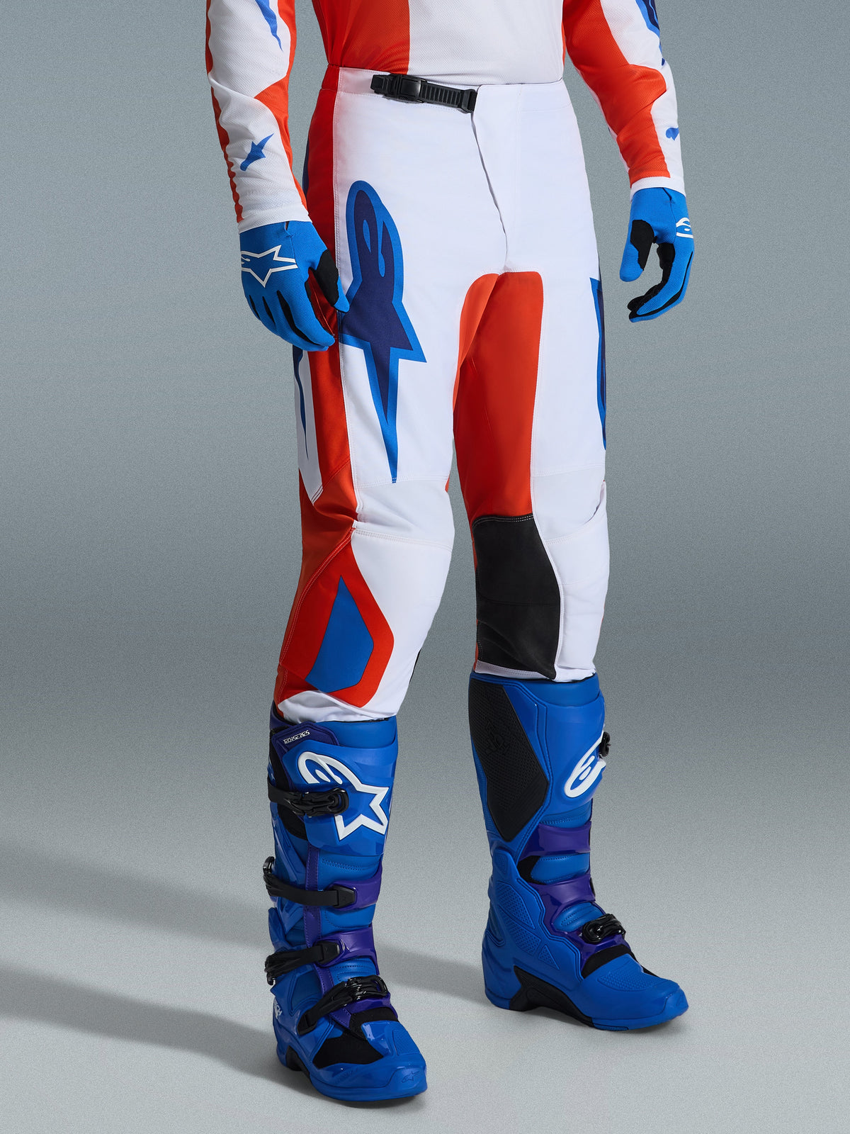 Alpinestars 2026 Fluid Grid Pants, spodnie motocrossowe, pomarańczowe i niebieskie UCLA, noszone z pasującymi niebieskimi rękawicami i butami, biała baza z wyrazistym graficznym brandingiem, czarne wewnętrzne panele na kolanach, ergonomiczny design do jazdy off-road