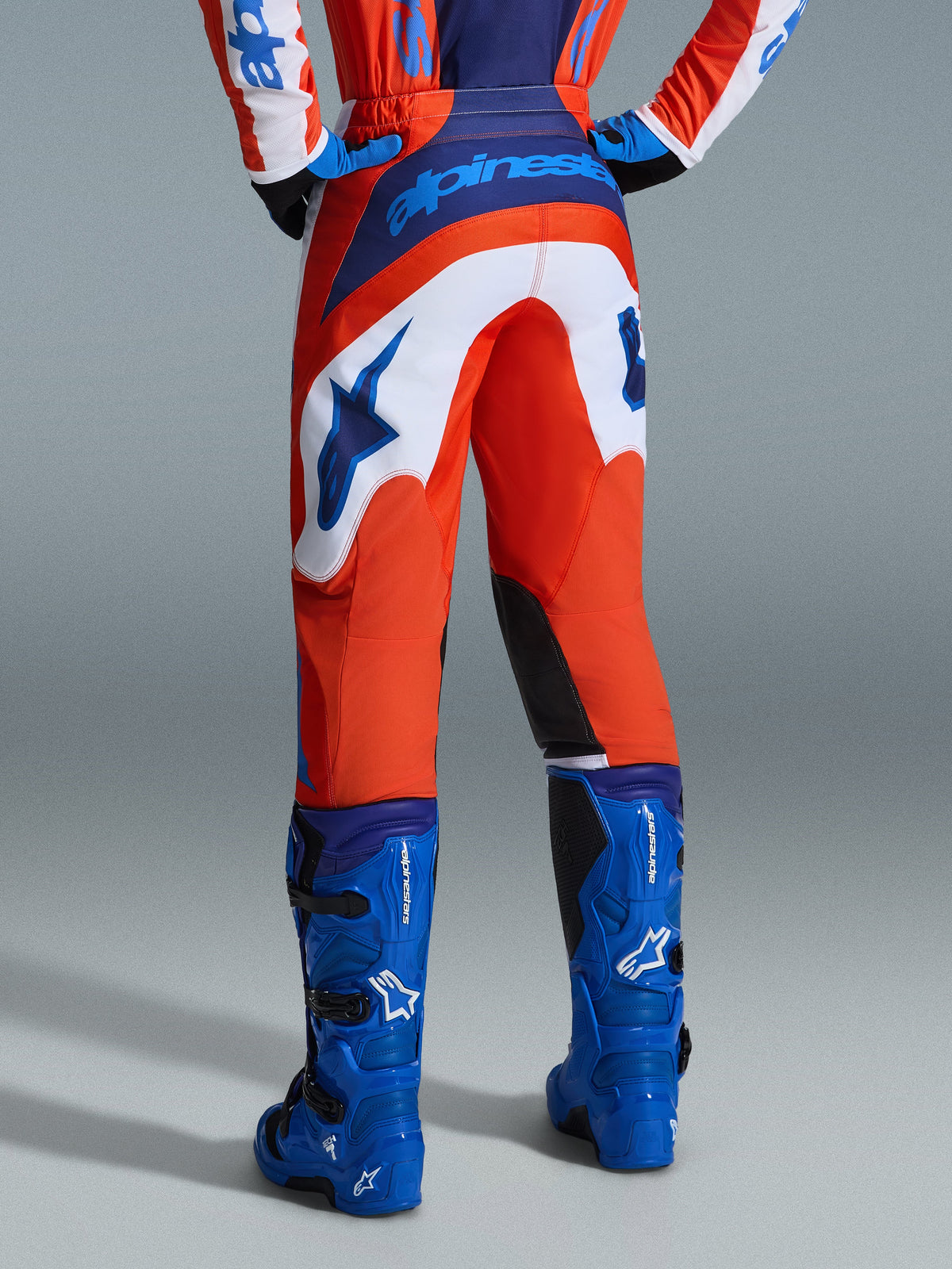 Alpinestars 2026 Fluid Grid Pants, spodnie motocrossowe, pomarańczowe i niebieskie UCLA z białymi panelami, widok z tyłu pokazujący wyraźne niebieskie logo na siedzeniu i logo gwiazdy, zestawione z pasującymi niebieskimi butami motocrossowymi, profesjonalny sprzęt do wyścigów off-road