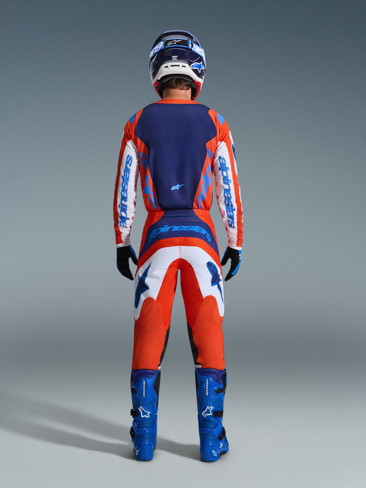Alpinestars 2026 Fluid Grid Pants, spodnie motocrossowe, pomarańczowe i niebieskie UCLA Blue, pełny widok od tyłu na motocyklistę w kompletnym zestawie odzieży wyścigowej z pasującą koszulką, niebieskimi butami i kaskiem, z wyraźnym brandingiem i ergonomicznymi panelami do jazdy w terenie.