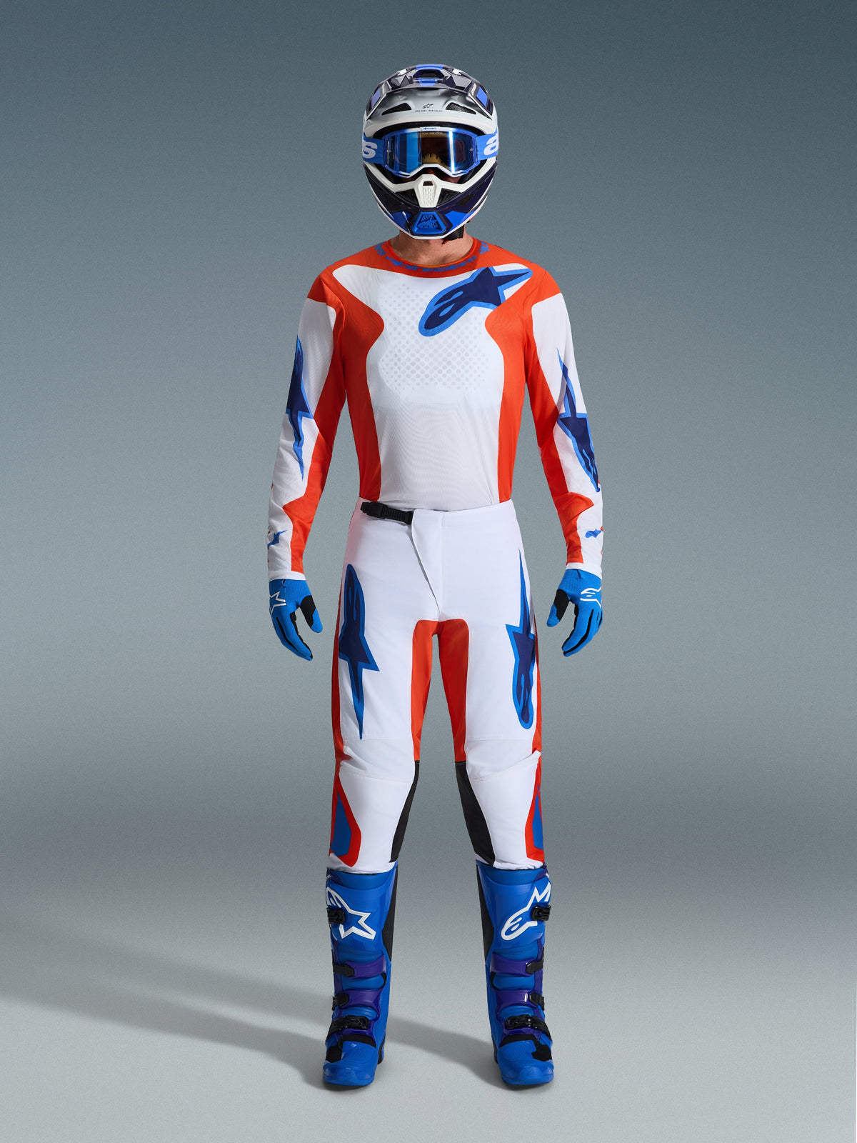 Zawodnik motocrossowy w pełnym stroju wyścigowym, spodnie Alpinestars 2026 Fluid Grid w kolorze pomarańczowym i UCLA Blue, biała koszulka z pomarańczowymi rękawami i niebieskim logo, dopasowany niebieski kask i gogle, niebieskie buty i niebieskie rękawice, skoordynowany zestaw do wyścigów off-road