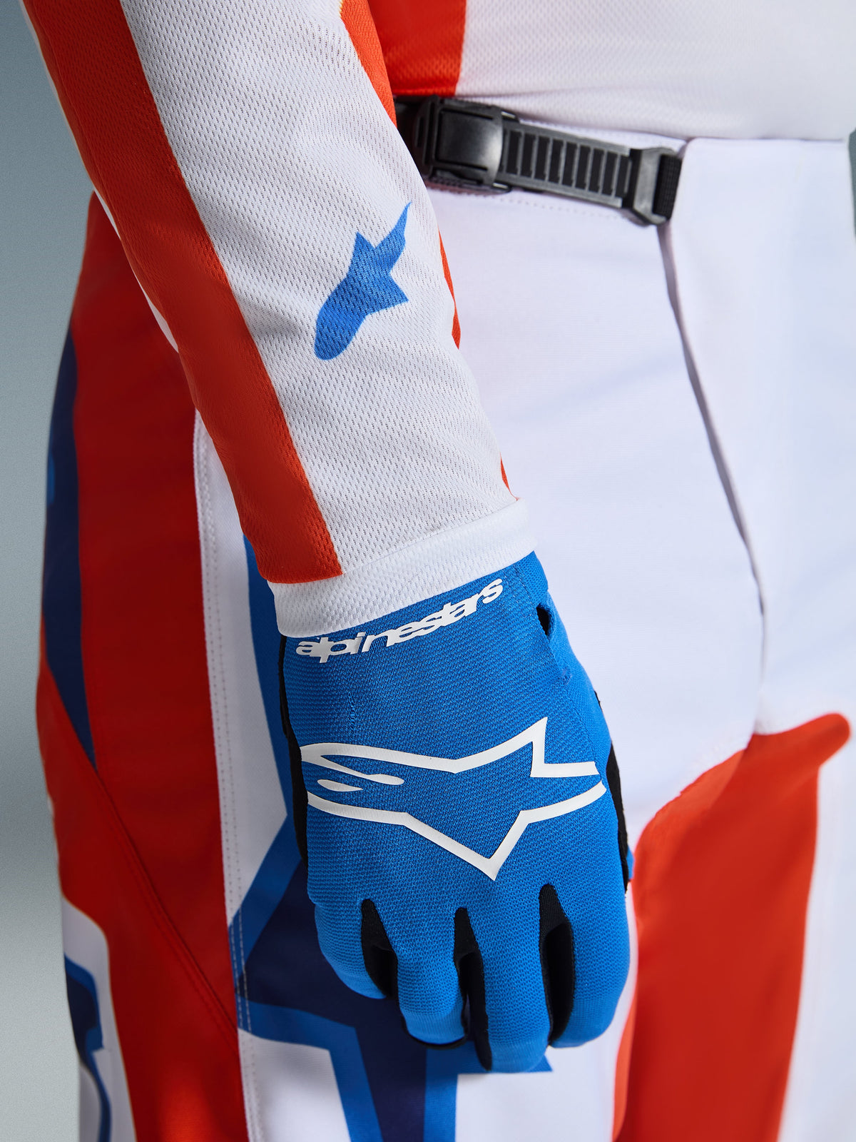 Spodnie Alpinestars 2026 Fluid Grid, spodnie motocrossowe, pomarańczowe i niebieskie (UCLA Blue), zbliżenie na dopasowaną niebieską rękawicę i biały rękaw koszulki z siatki z niebieskimi logotypami, widoczne zapięcie na zapadkę w pasie i sportowy krój.