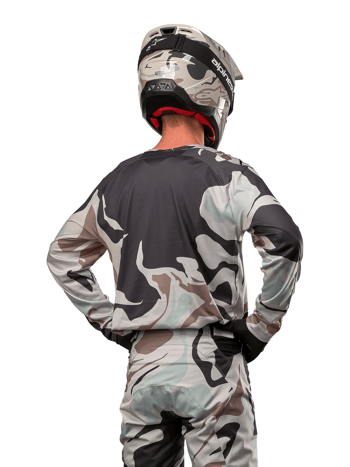 Zawodnik motocrossowy widziany od tyłu w koszulce Alpinestars 2024 Racer Tactical w kolorze Iron Camo Dust Gray, pasujące spodnie i kask z motywem kamuflażu, ręce na biodrach, dopasowany strój do wyścigów terenowych.