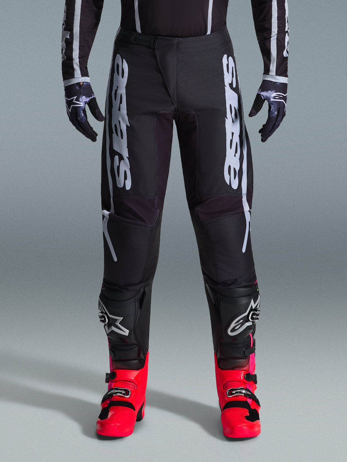 Alpinestars 2026 Fluid Apex Pants, czarne spodnie motocrossowe z dużymi białymi napisami, noszone z czarnymi ochraniaczami kolan i jaskrawoczerwonymi butami, lekka konstrukcja z materiału poly-fabric 600D, sportowy krój do wyścigów off-road