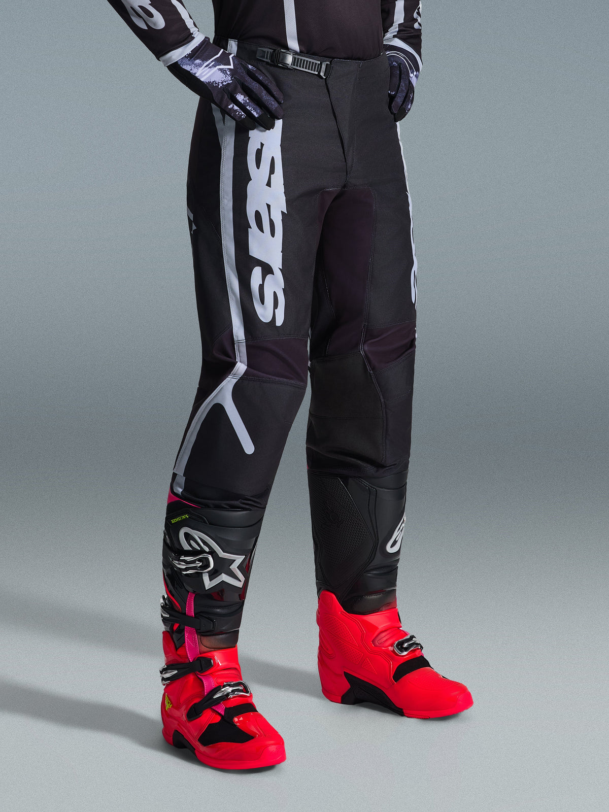 Spodnie Alpinestars Fluid Apex 2026, spodnie motocrossowe, czarno-szare, noszone z jaskraworóżowymi i czerwonymi butami oraz pasującą czarno-białą bluzą, wyraźny pionowy napis Alpinestars na udzie, wzmocnione panele i zapięcie na zapadkę w pasie, wysoka wydajność off-road