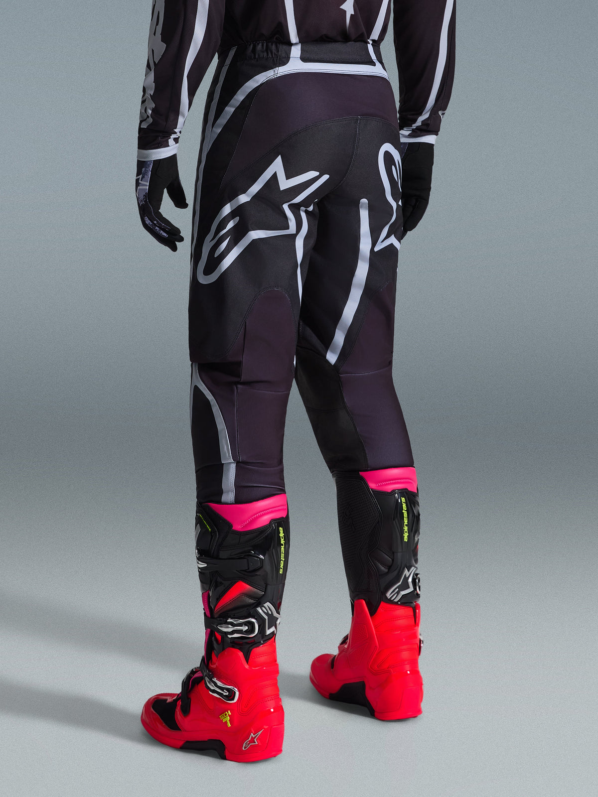 Spodnie Alpinestars 2026 Fluid Apex, spodnie motocrossowe, czarno-szare, widok z tyłu z dużymi białymi logotypami Alpinestars na siedzeniu, noszone z jaskrawoczerwonymi i różowymi butami, wytrzymała konstrukcja z materiału polyfabric 600D zapewniająca wydajność off-road