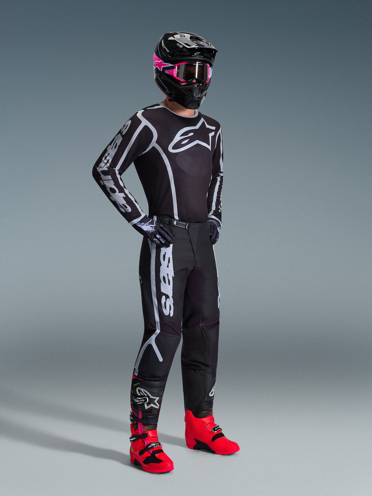 Alpinestars 2026 Fluid Apex Pants, spodnie motocrossowe, czarno-szare, zestawione z pasującą czarno-białą koszulką, czarnym kaskiem z różowymi goglami i jaskrawoczerwonymi butami, z wyraźnym białym logo na nogawkach i sportowym krojem do jazdy terenowej.