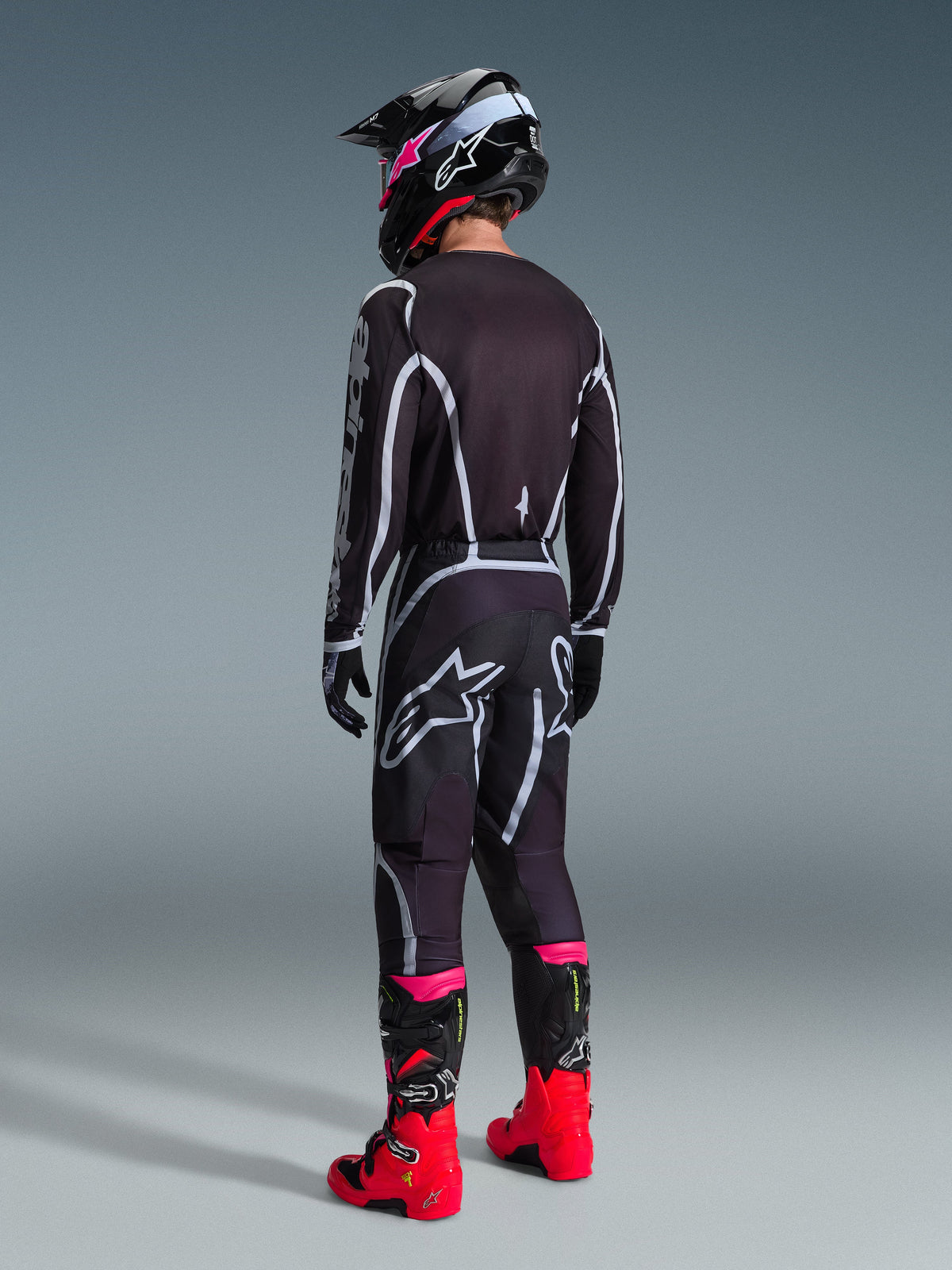 Alpinestars 2026 Fluid Apex Pants, spodnie motocrossowe, czarno-szare, widok z tyłu z białym logo Alpinestars na siedzeniu, noszone z pasującą koszulką, czarno-różowym kaskiem i jaskrawoczerwonymi butami.