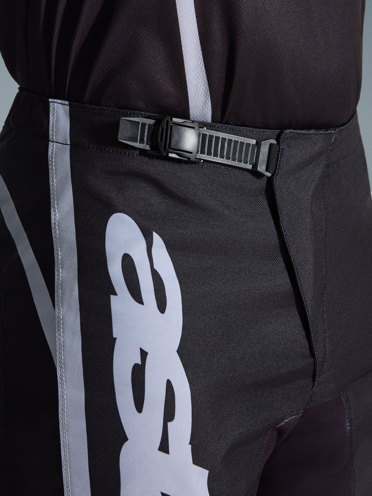 Alpinestars 2026 Fluid Apex Pants, spodnie motocrossowe, czarno-szare, zbliżenie na zintegrowany system zapięcia zapadkowego zapewniający pewne dopasowanie, biała grafika logo na wytrzymałym poliestrze 600D.
