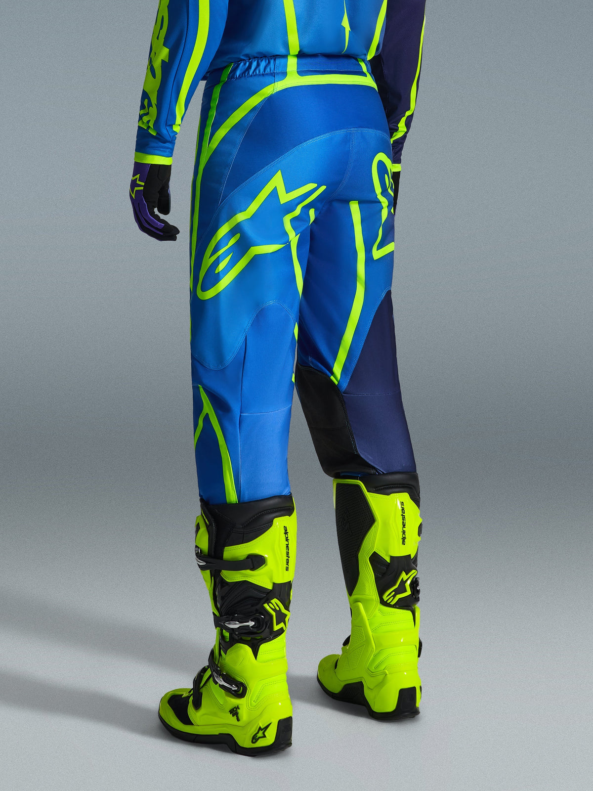 Alpinestars 2026 Fluid Apex Pants, spodnie motocrossowe, niebieskie i żółte fluo, widok od tyłu na motocyklistę w pasującym stroju i jaskrawożółtych butach, wyraziste logo Alpinestars, ergonomiczny design