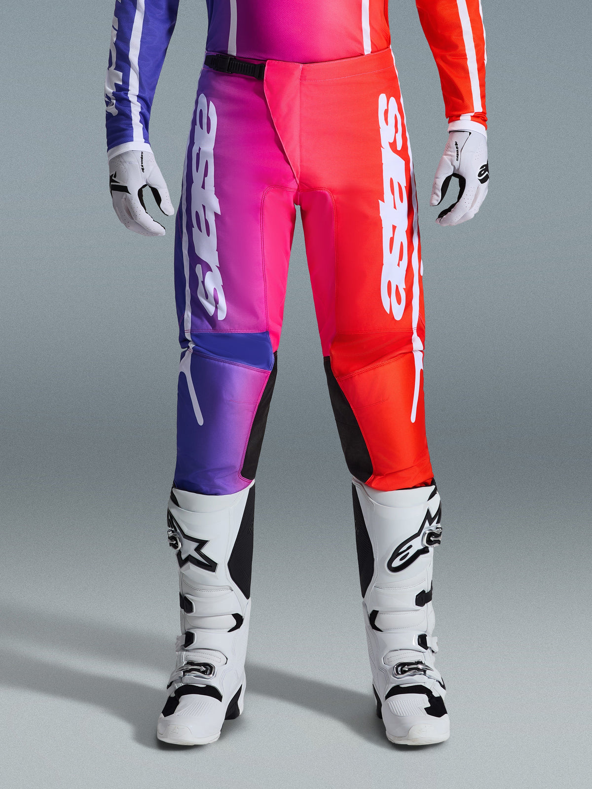 Alpinestars 2026 Fluid Apex Pants, spodnie motocrossowe, wielokolorowy wzór z gradientem fioletu, różu i czerwieni, duże białe logo Alpinestars na nogawkach, zestawione z biało-czarnymi butami i rękawicami motocrossowymi, profesjonalny sprzęt do wyścigów terenowych