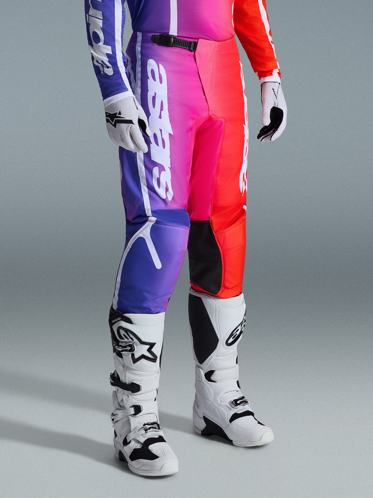 Alpinestars 2026 Fluid Apex Pants, spodnie motocrossowe, wielokolorowy gradient fioletu, różu i czerwieni, zestawione z biało-czarnymi butami i rękawicami motocrossowymi, z białym pionowym logo i czarnymi akcentami na kolanach, profesjonalna odzież off-road
