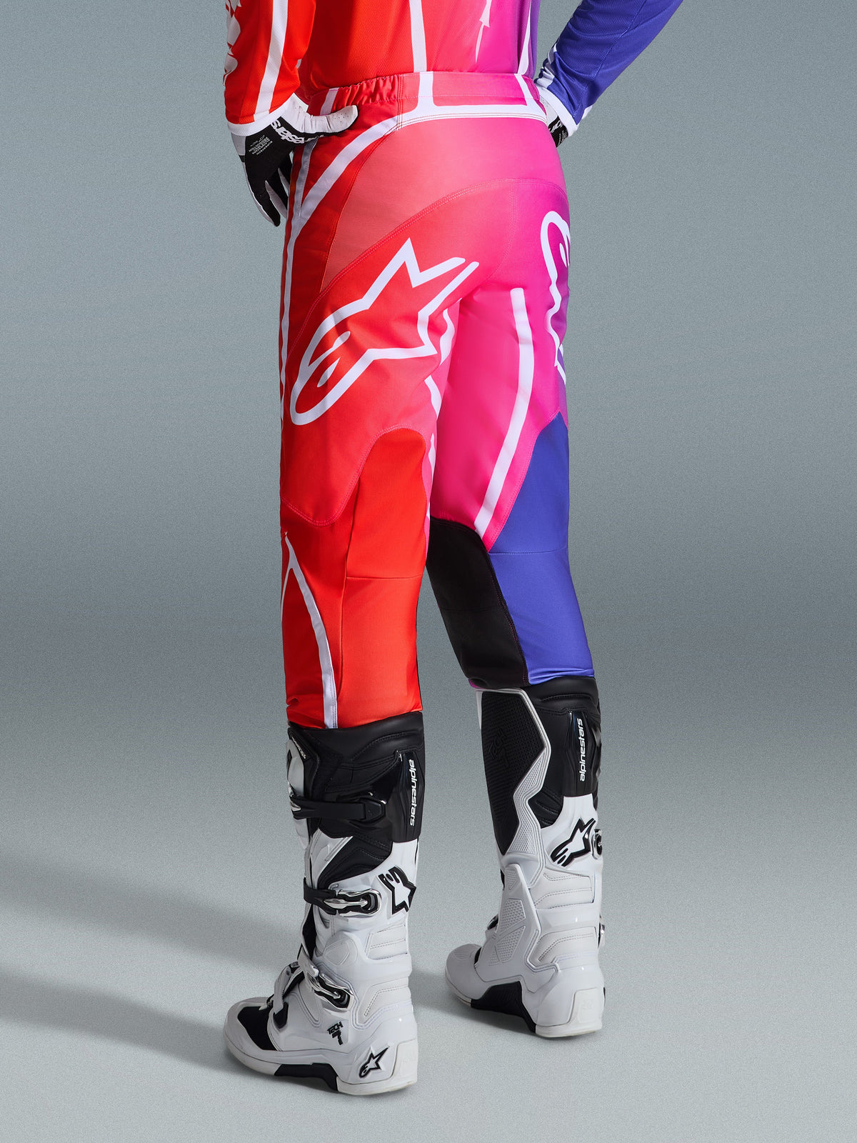 Alpinestars 2026 Fluid Apex Pants, spodnie motocrossowe, wielokolorowe czerwone, różowe i fioletowe, widok z tyłu na motocyklistę w spodniach i biało-czarnych butach motocrossowych, duże białe logo Alpinestars, jaskrawy gradientowy wzór