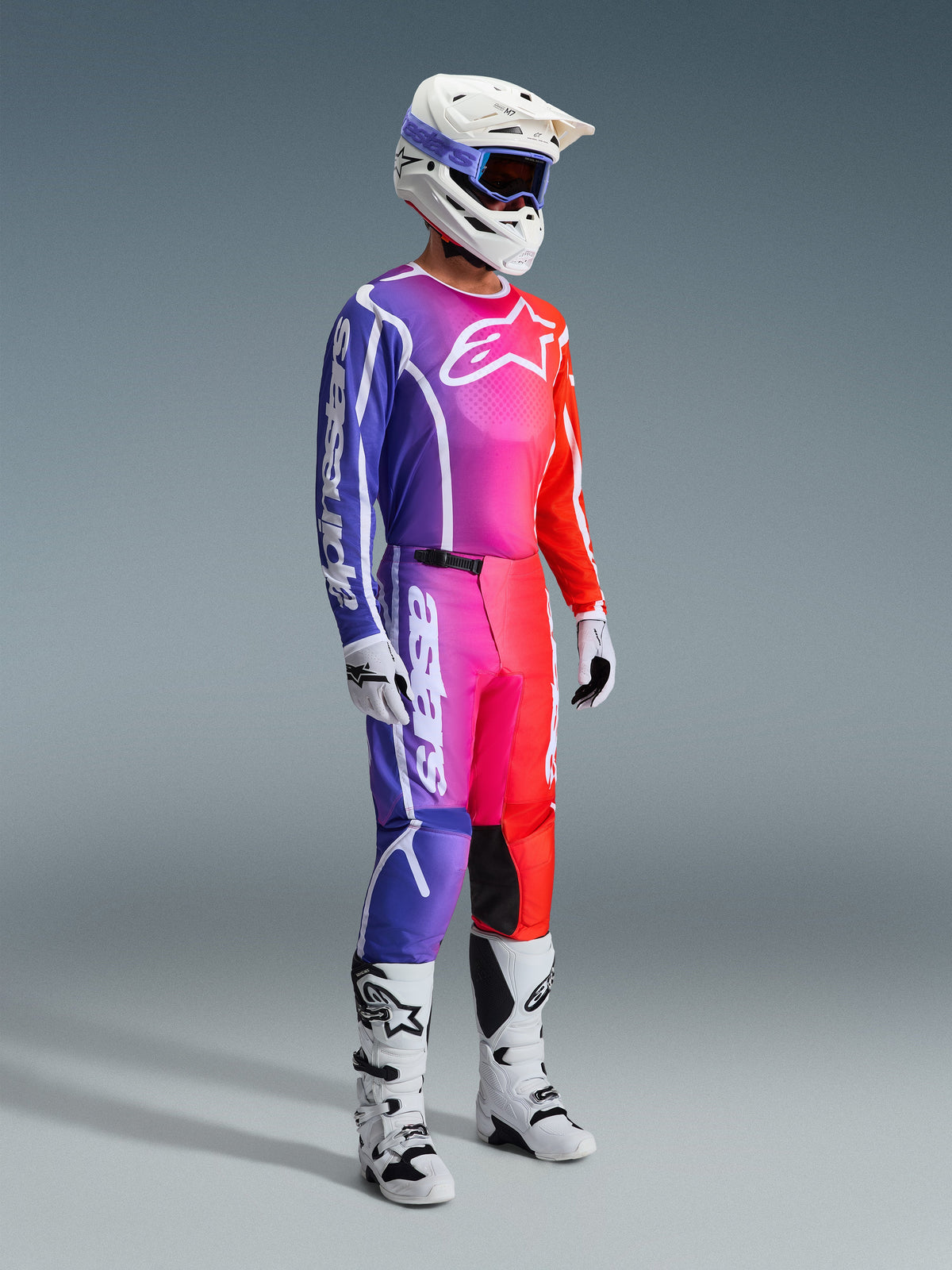 Alpinestars 2026 Fluid Apex Pants, spodnie motocrossowe, wielokolorowy gradient fioletu, różu i pomarańczu, zestawione z dopasowaną bluzą, białym kaskiem i białymi butami, z wyraźnym białym logo i ergonomicznym designem do wyścigów terenowych