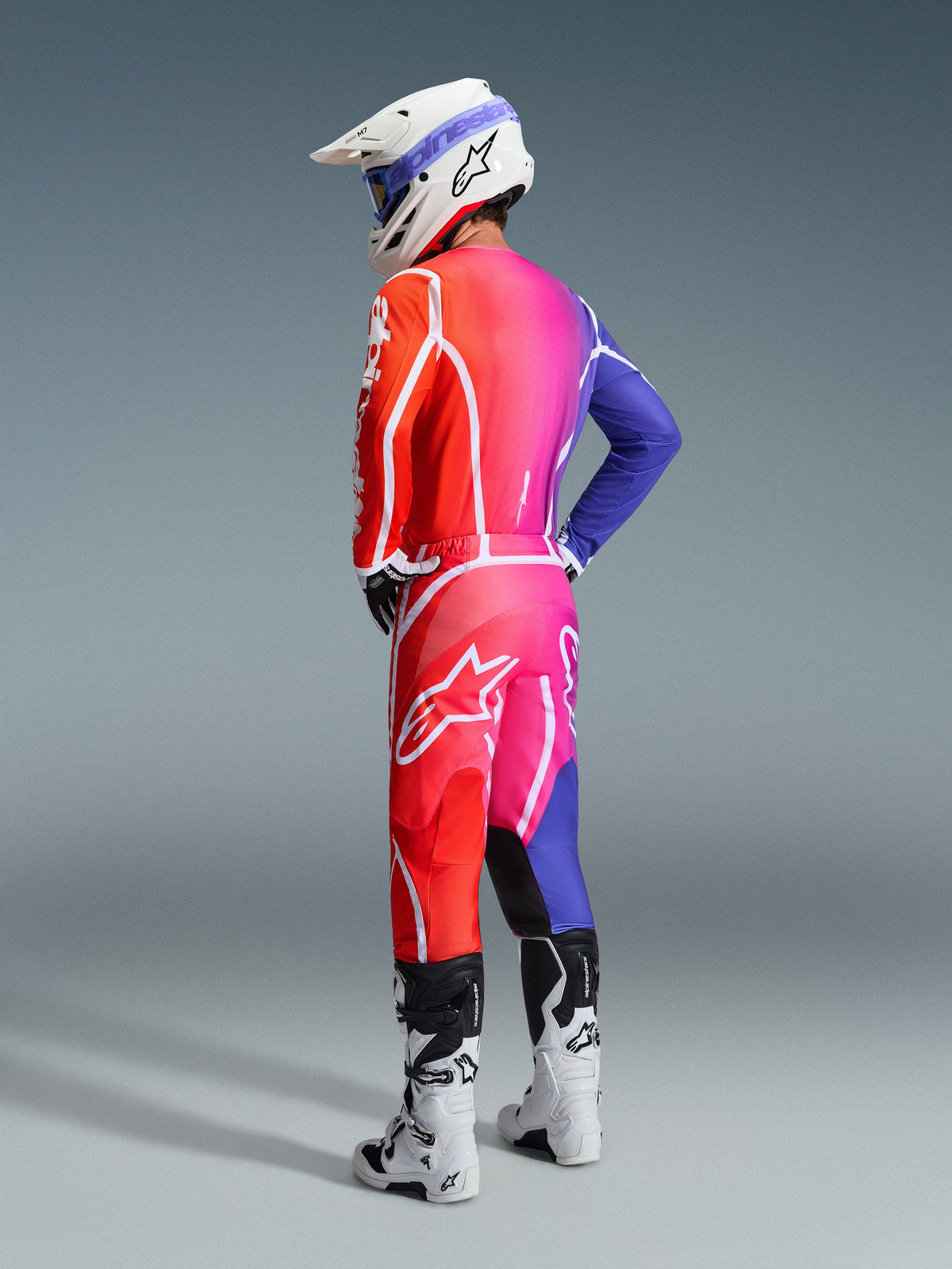 Zawodnik motocrossowy w stroju Alpinestars 2026 Fluid Apex, wielokolorowa bluza i spodnie z gradientem czerwieni, różu i fioletu, białe logo, biały kask i białe buty, dopasowany strój do wyścigów off-road