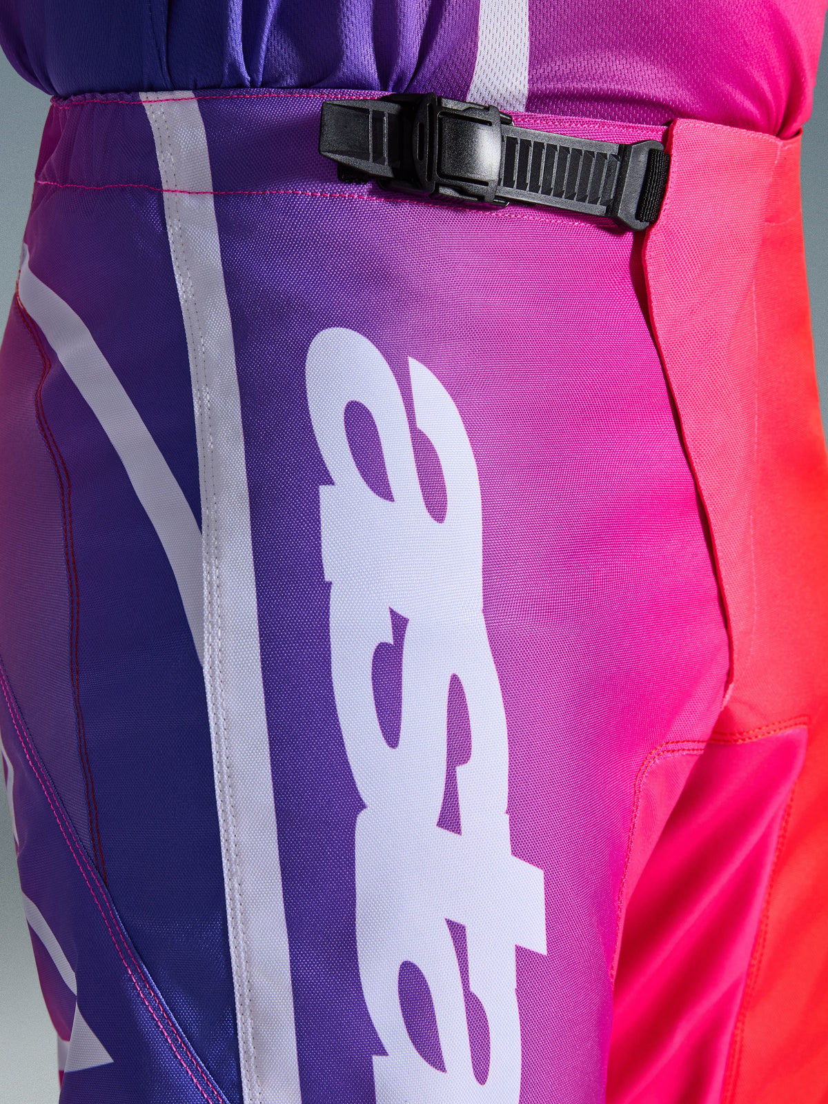Alpinestars 2026 Fluid Apex Pants, spodnie motocrossowe, wielokolorowe fioletowo-różowo-czerwone, zbliżenie na talię i biodra z czarnym systemem zapięcia zapadkowego i dużym białym pionowym logo Alpinestars, żywy gradientowy wzór z wytrzymałą teksturą materiału