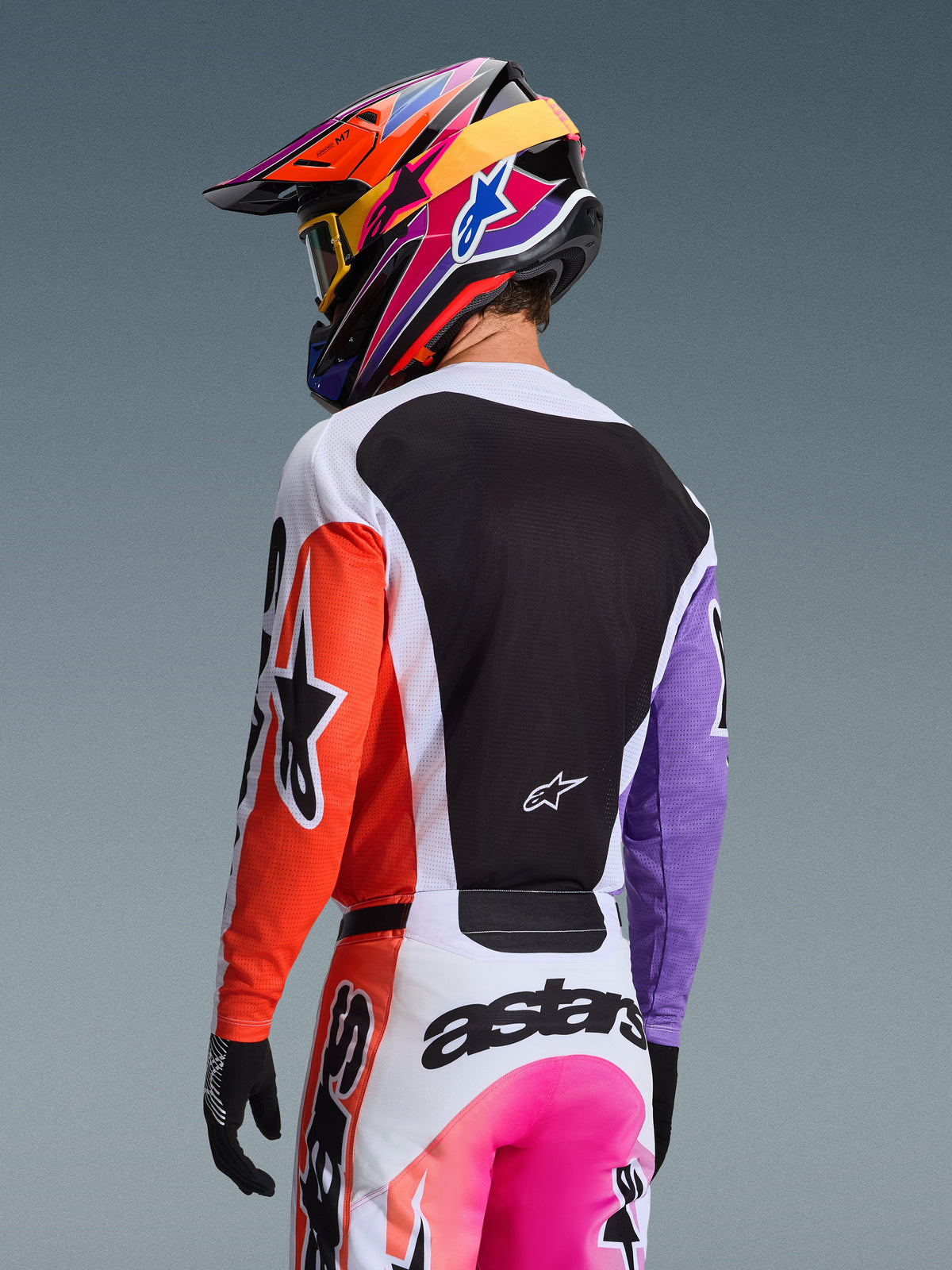 Motocrossista widoczny od tyłu w koszulce Alpinestars 2026 Racer Air Portl Multicolor, pomarańczowy prawy rękaw, fioletowy lewy rękaw, duży czarny panel z siatki na plecach zapewniający wentylację, dopasowane spodnie motocrossowe i wielokolorowy kask, profesjonalny sprzęt do wyścigów off-road