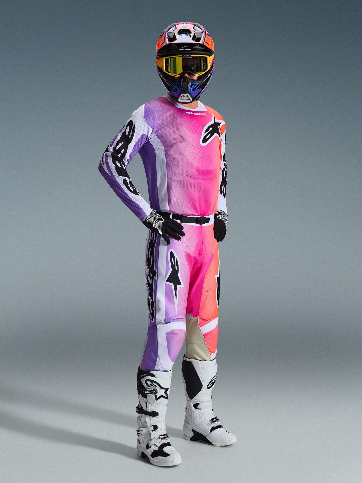 Zawodnik motocrossowy w koszulce Alpinestars 2026 Racer Air Portl i pasujących spodniach, żywy gradient w kolorach różowym, fioletowym i pomarańczowym z białymi akcentami, dopasowany kolorowy kask i żółte gogle, biało-czarne buty, kompletny strój wyścigowy do jazdy off-road