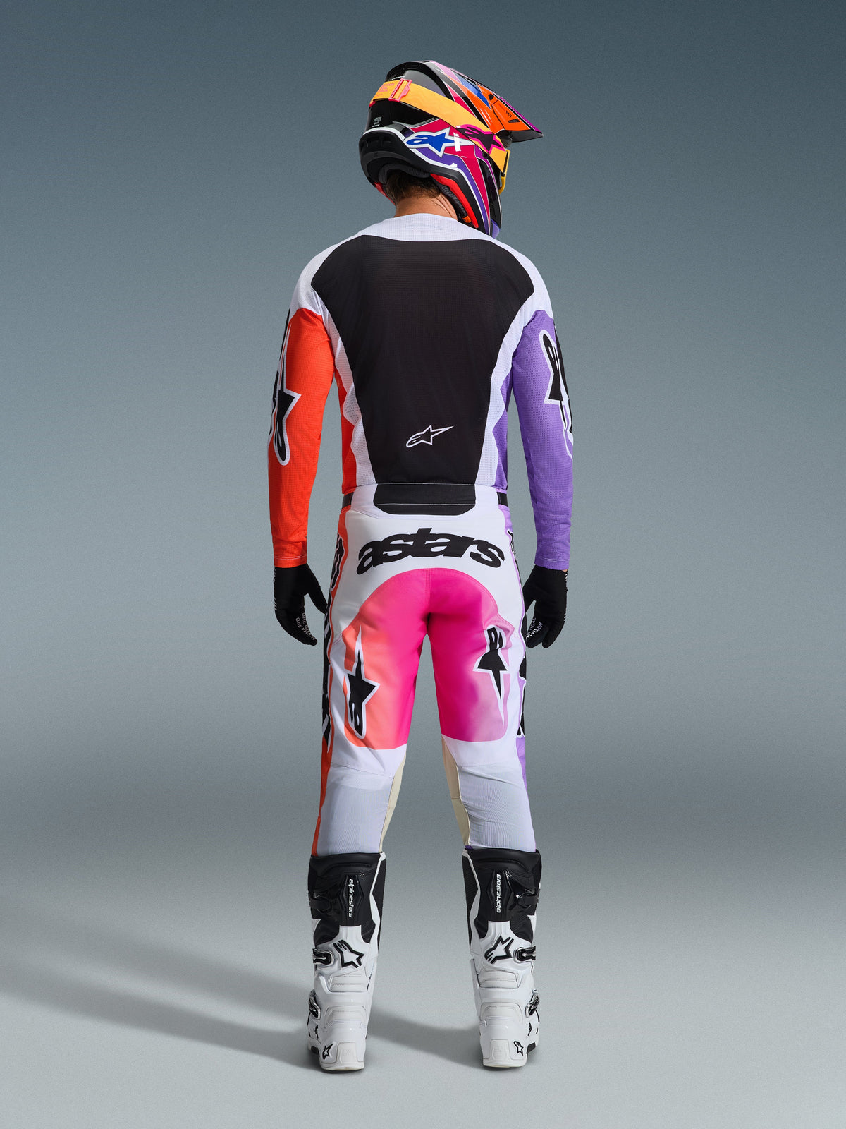 Zawodnik motocrossowy w pełnym stroju wyścigowym widoczny od tyłu, koszulka Alpinestars 2026 Racer Air Portl z białymi, czarnymi, pomarańczowymi i fioletowymi panelami, białe spodnie z różowymi akcentami i brandingiem „astars”, biało-czarne buty oraz wielokolorowy kask, skoordynowany zestaw do wyścigów motocrossowych