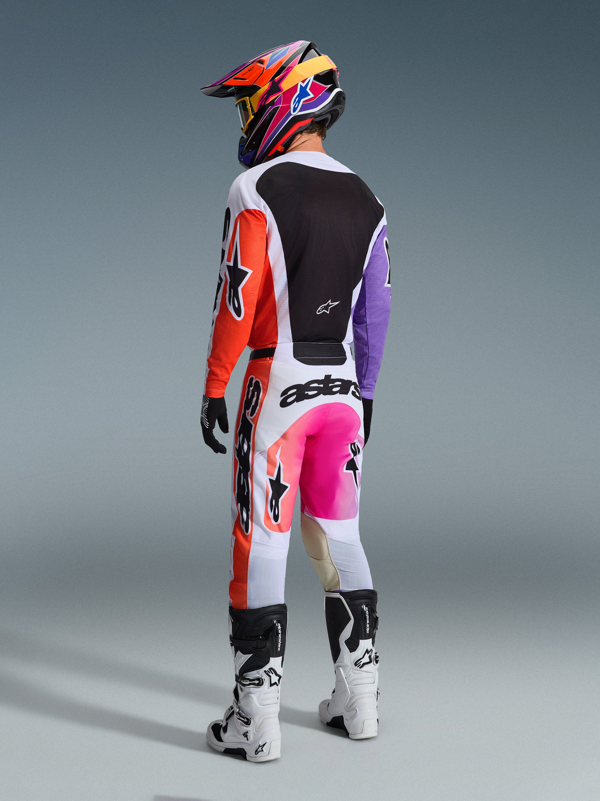 Alpinestars 2026 Racer Air Portl Jersey, wielokolorowa koszulka motocrossowa, widok z tyłu na motocyklistę w pełnym rynsztunku, biała baza z pomarańczowymi i fioletowymi rękawami oraz czarnym siatkowym panelem na plecach, w zestawieniu z dopasowanymi różowo-pomarańczowymi spodniami, kolorowym kaskiem i białymi butami, profesjonalny strój do wyścigów terenowych