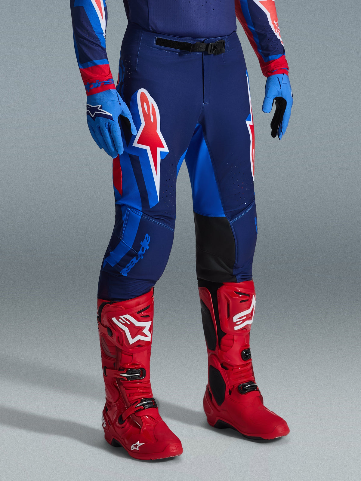 Alpinestars 2026 Supertech Vista Pants, spodnie motocrossowe, kolorystyka Dark Navy Hot Coral Cobalt, zestawione z jaskrawoczerwonymi butami wyścigowymi i niebieskimi rękawicami, duże graficzne logo na udach i czarne wzmocnione panele po wewnętrznej stronie kolan dla wydajności w terenie.