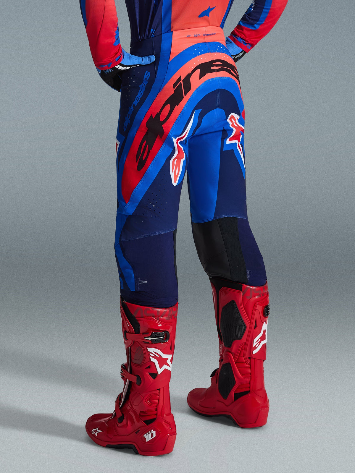 Alpinestars 2026 Supertech Vista Pants, spodnie motocrossowe, kolor granatowy, koralowy i kobaltowy, widok zawodnika z tyłu, w zestawieniu z czerwonymi butami motocrossowymi, wyraźne logo na siedzeniu, sportowy krój ze strefami elastycznymi i wytrzymała konstrukcja rip-stop do profesjonalnych wyścigów