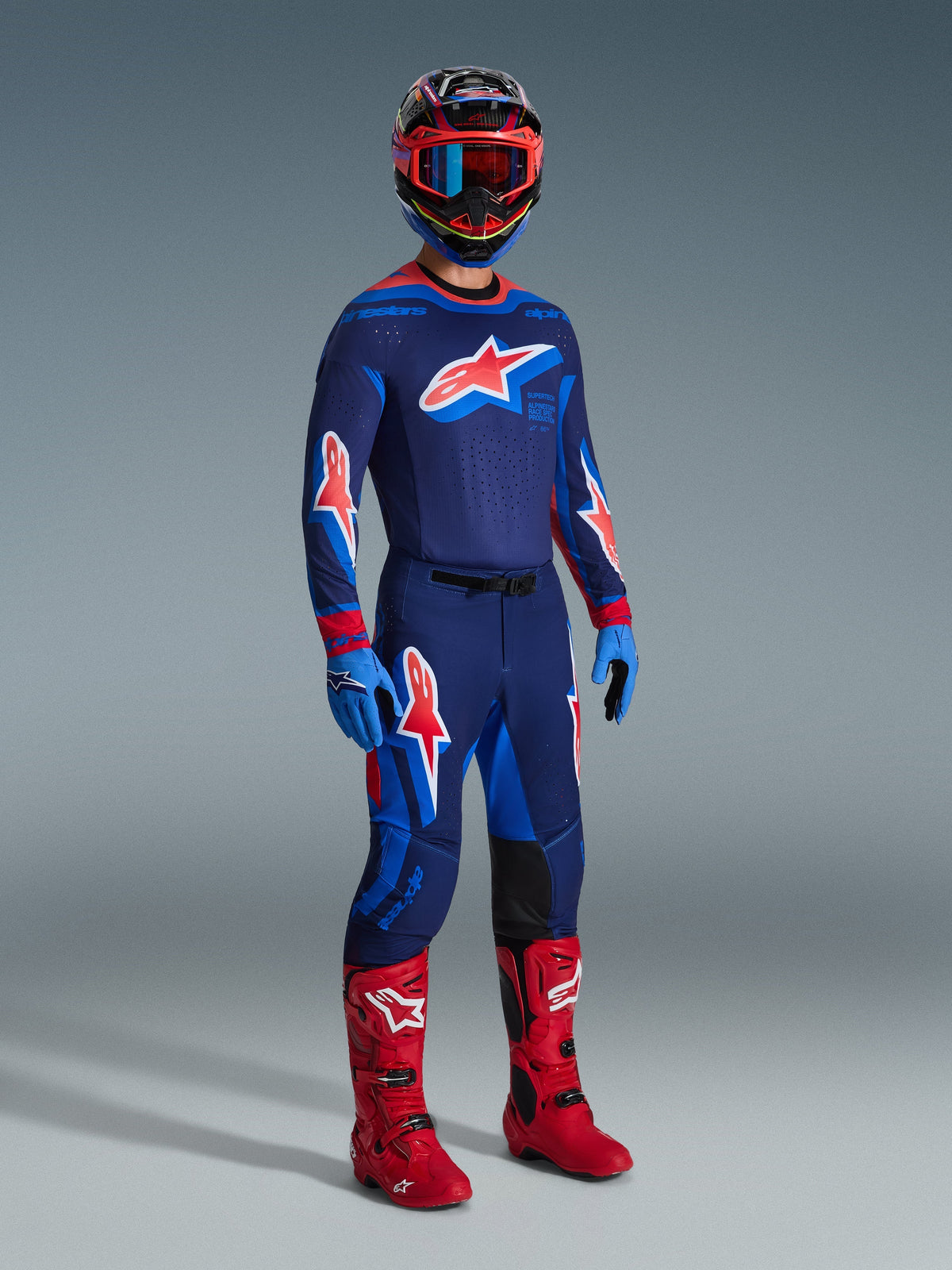 Motocyklista motocrossowy w spodniach 2026 Supertech Vista w kolorze Dark Navy Hot Coral Cobalt, dopasowana koszulka wyścigowa z długim rękawem, czerwone buty motocrossowe i kask z czerwonymi goglami, ujęcie studyjne całej sylwetki profesjonalnego stroju off-road