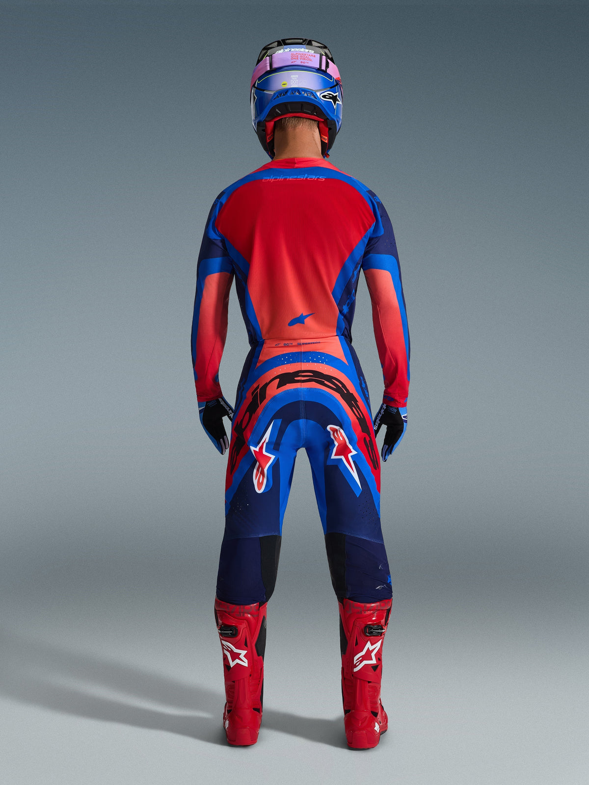 Alpinestars 2026 Supertech Vista Pants, spodnie motocrossowe, kolory Dark Navy, Hot Coral i Cobalt, widok z tyłu na motocyklistę w pełnym stroju wyścigowym z dopasowaną koszulką, niebieskim kaskiem i czerwonymi butami, widoczny wyraźny branding na siedzeniu i nogawkach.