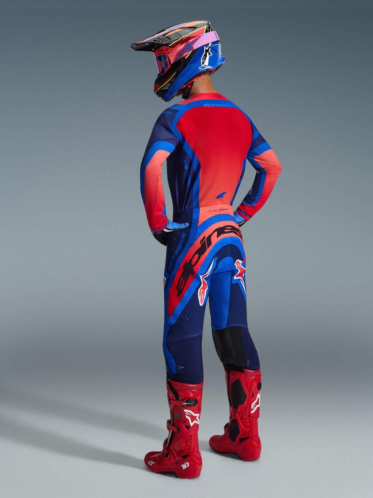 Alpinestars 2026 Supertech Vista Pants, spodnie motocrossowe, kolorystyka Dark Navy Hot Coral i Cobalt, widok z tyłu na motocyklistę w pasującej bluzie i czerwonych butach, wyraźne logo na siedzeniu, wytrzymały elastyczny materiał.