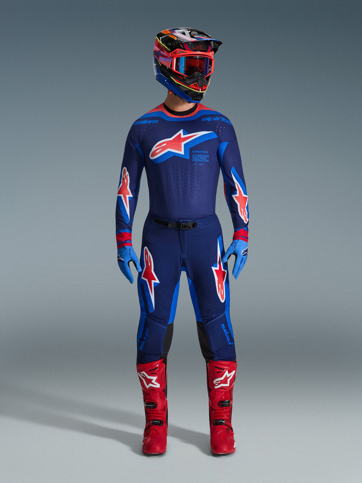 Zawodnik motocrossowy w kompletnym stroju wyścigowym, spodnie Alpinestars Supertech Vista 2026 w kolorystyce Dark Navy Hot Coral Cobalt, dopasowana koszulka z wyraźnym logo gwiazdy, jaskrawoczerwone buty oraz kask z czerwonymi goglami, profesjonalny zestaw off-road