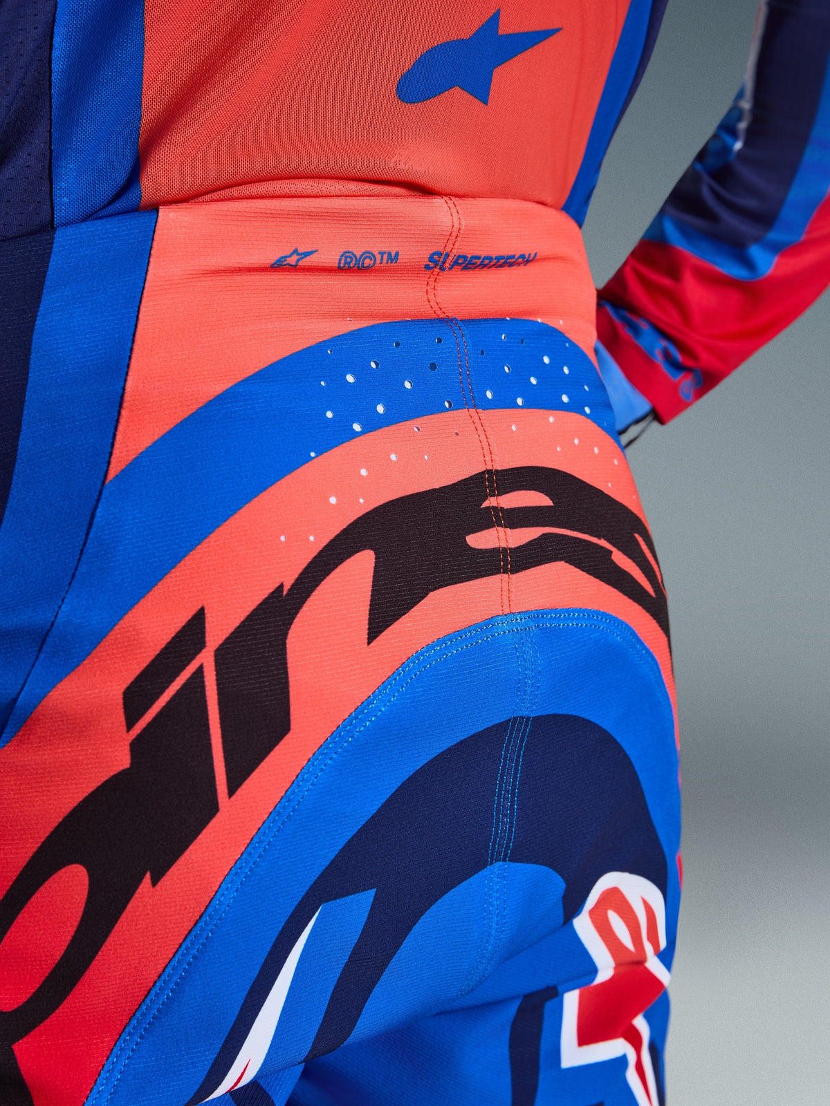 Zbliżenie na spodnie Alpinestars 2026 Supertech Vista, spodnie motocrossowe, w kolorach jaskrawego koralu i kobaltu, z dużą czarną grafiką Alpinestars na siedzeniu, laserowo wycinanymi otworami wentylacyjnymi i brandingiem „SUPERTECH” z tyłu w pasie, profesjonalna odzież wyścigowa.