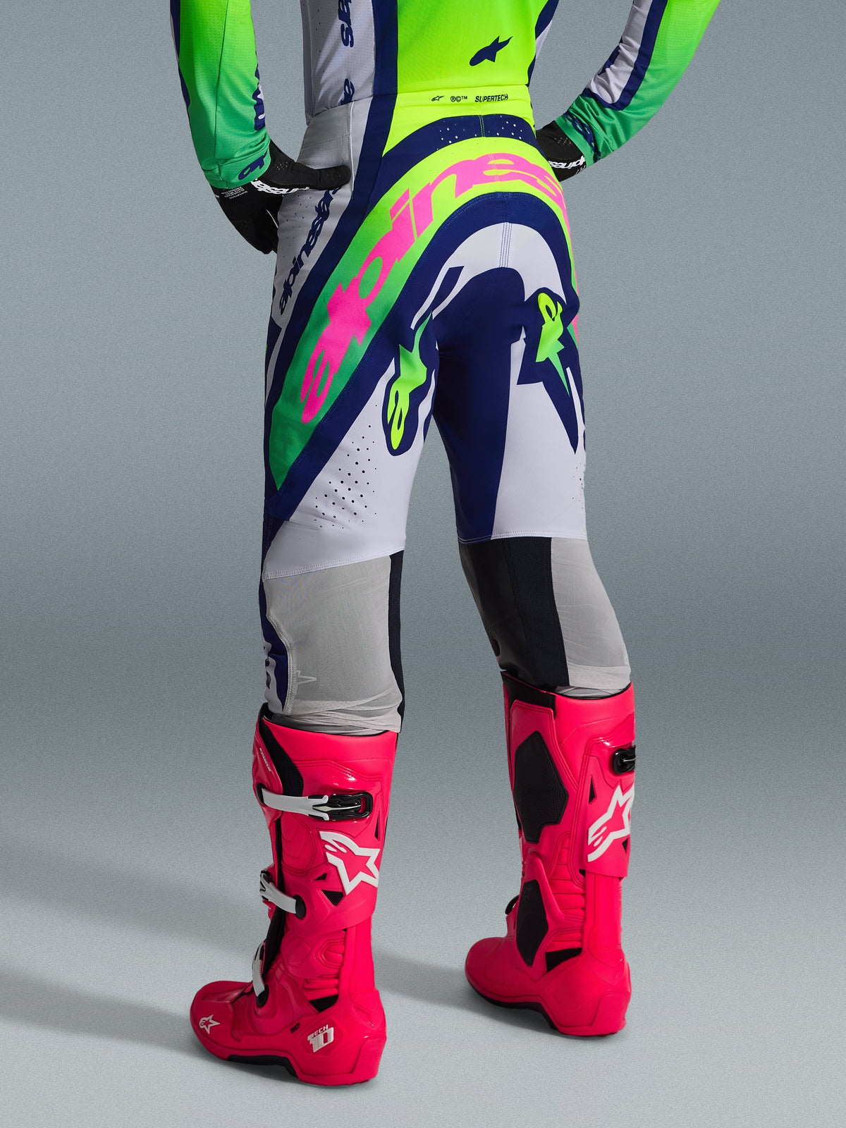 Alpinestars 2026 Supertech Vista Pants, spodnie motocrossowe, jasnoszare, żółte fluo i niebieskie, widok z tyłu z dużym różowym logo Alpinestars, noszone z pasującą koszulką wyścigową i jaskraworóżowymi butami Tech 10, profesjonalna odzież off-road