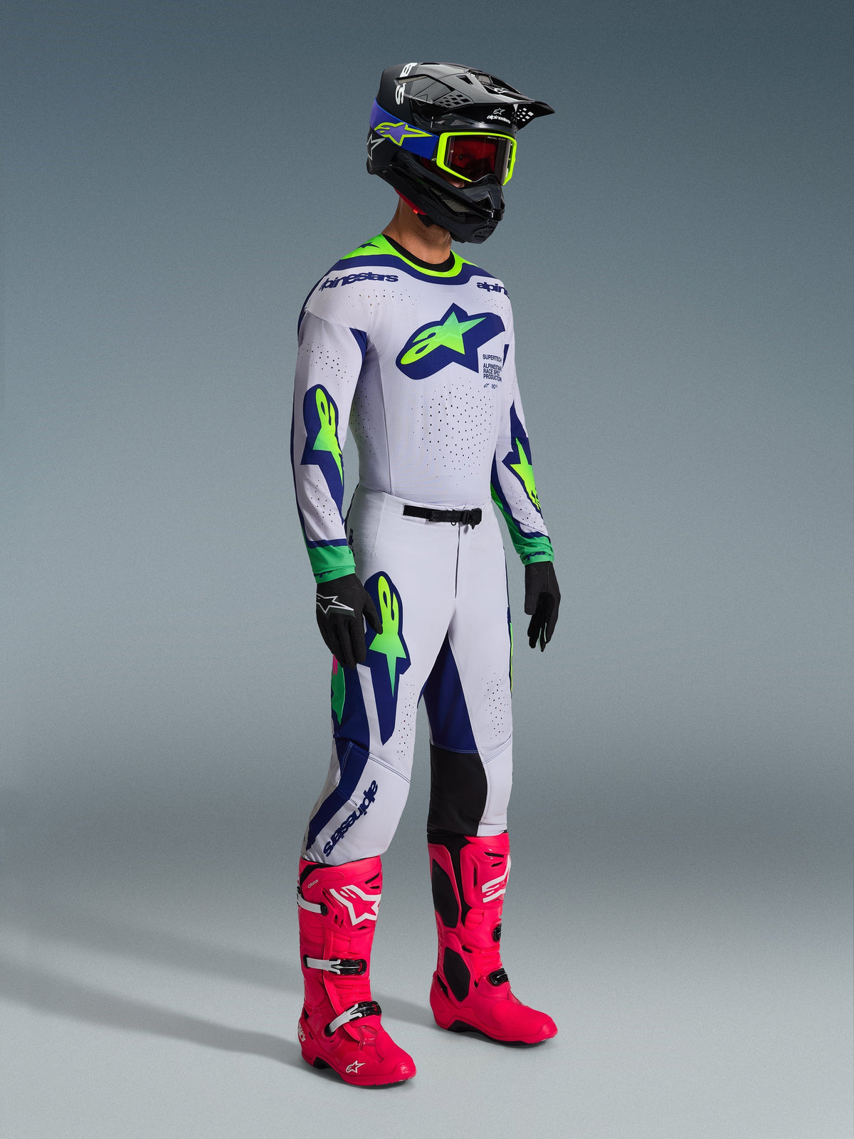 Spodnie i dopasowana koszulka Alpinestars 2026 Supertech Vista, strój motocrossowy w kolorze jasnoszarym z żółtymi akcentami fluo i niebieskimi detalami, noszony przez motocyklistę w czarnym kasku i jaskraworóżowych butach, z wyrazistą sportową grafiką i profesjonalnym krojem wyścigowym