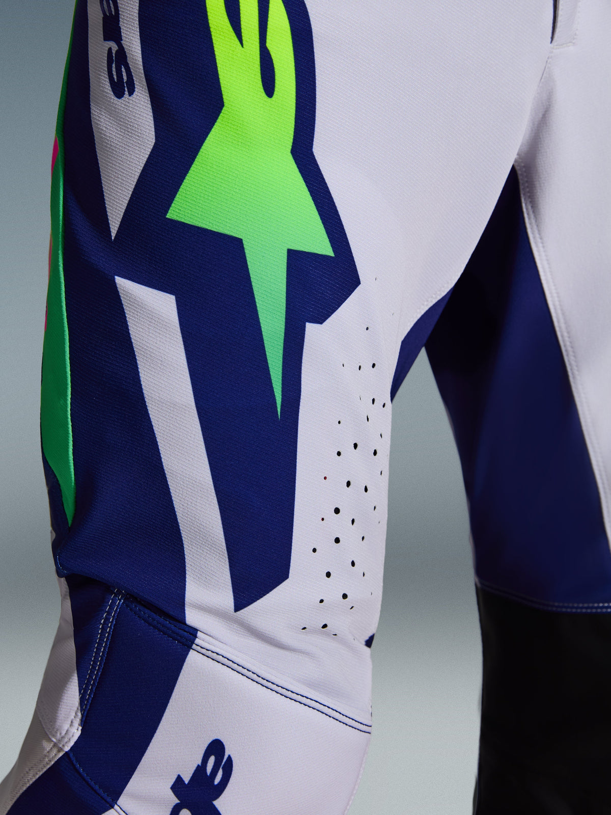 Alpinestars 2026 Supertech Vista Pants, spodnie motocrossowe, jasnoszare, żółte fluo i niebieskie, zbliżenie na udo i kolano, neonowe zielone logo gwiazdy na niebieskim panelu, laserowo wycinane perforacje wentylacyjne, czarne panele wewnętrzne na kolanach.