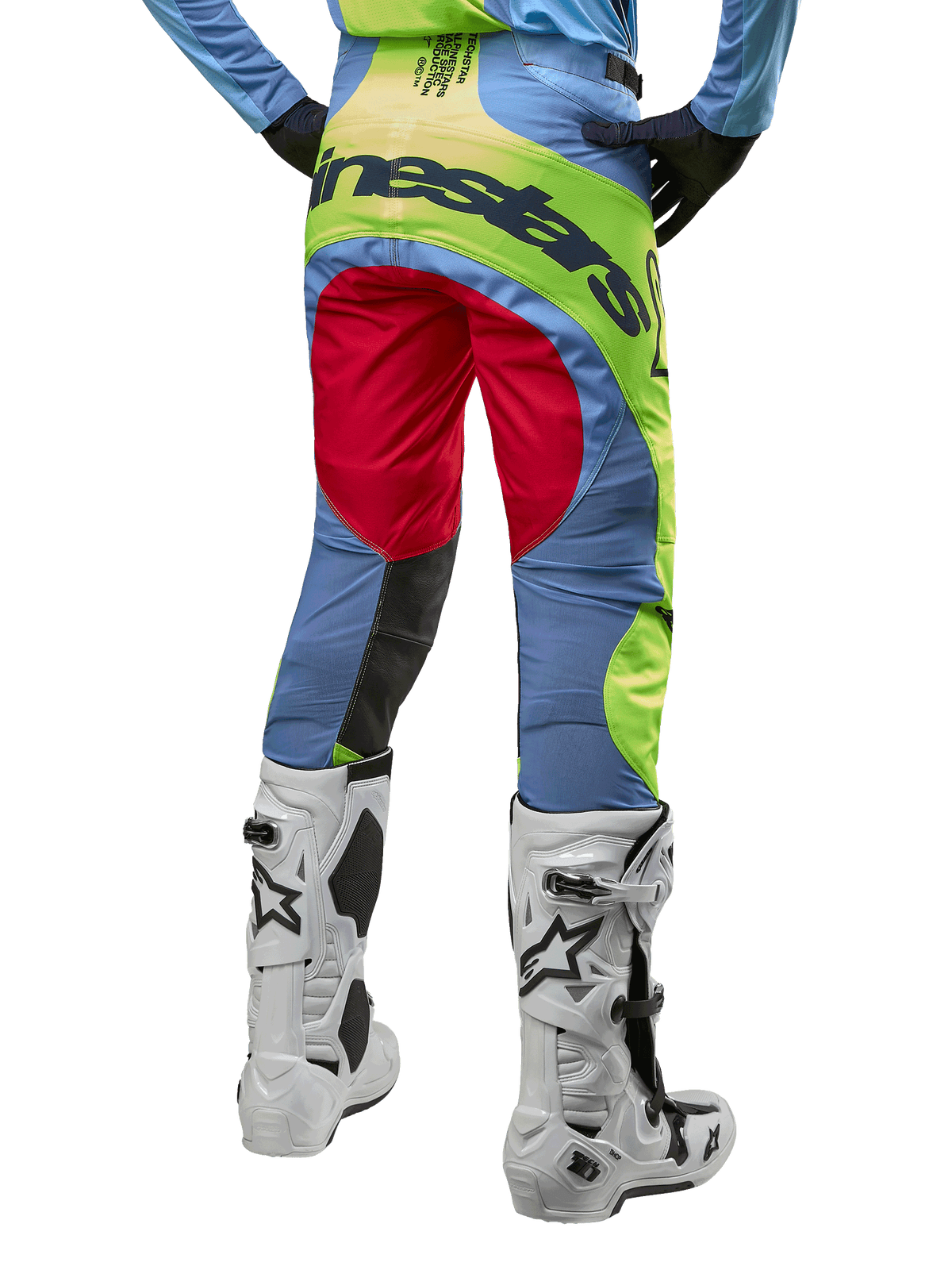 Alpinestars Techstar Ocuri Pants 2024, spodnie motocrossowe, jasnoniebieskie, neonowe żółte i czerwone, widok z tyłu w biało-czarnych butach motocrossowych, wyrazisty design color-blocking i duże logo Alpinestars na siedzeniu