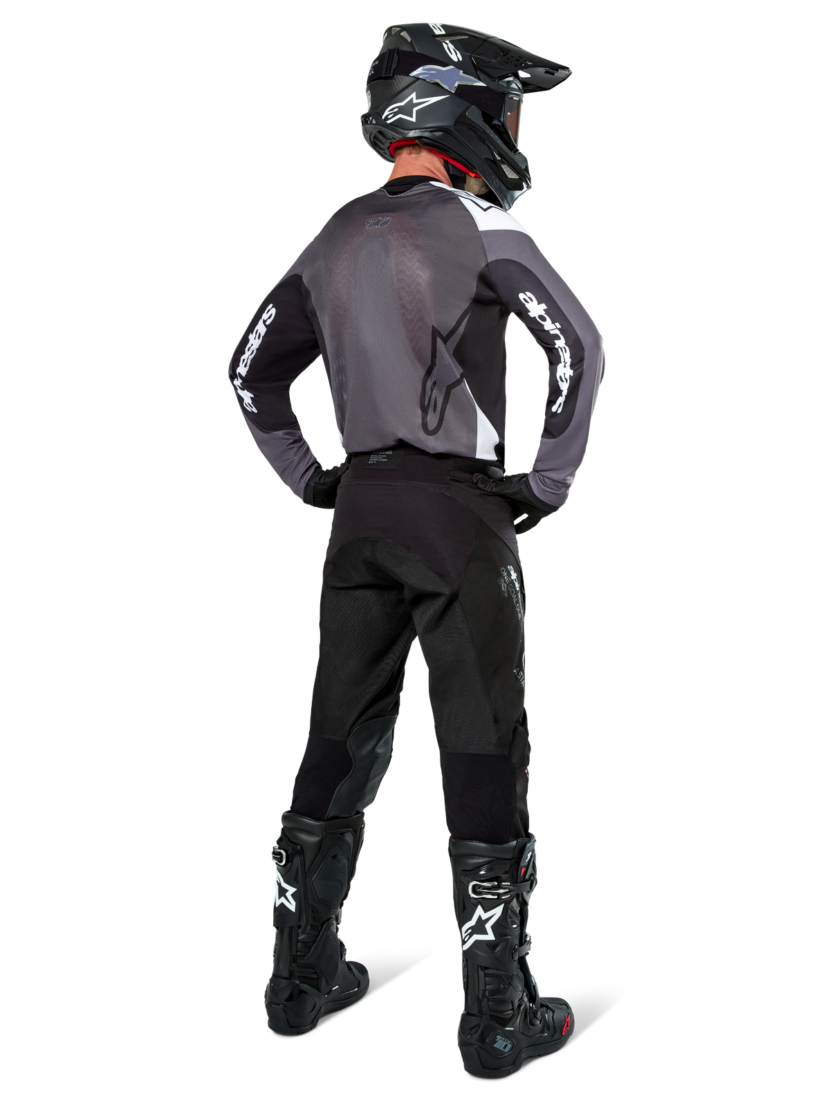 Zawodnik motocrossowy w koszulce Alpinestars Pro-Dura w kolorze Black Magnet White, widok z tyłu ukazujący szary siatkowy panel na plecach i czarne rękawy z białym brandingiem Alpinestars, zestawiony z czarnymi spodniami, czarnym kaskiem i czarno-białymi butami, profesjonalna odzież enduro