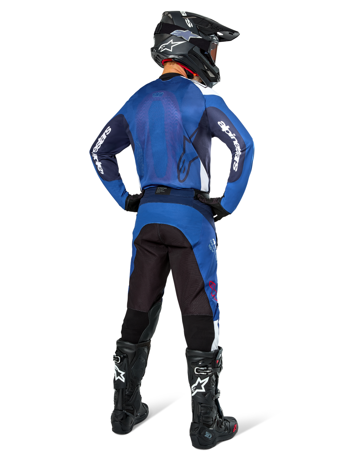 Motocyklista motocrossowy widoczny od tyłu w koszulce Alpinestars Pro-Dura w kolorze granatowym i jasnoniebieskim, pasujące niebieskie i czarne spodnie, czarny kask motocrossowy z goglami oraz czarne buty, prezentujący dopasowany strój do wyścigów terenowych