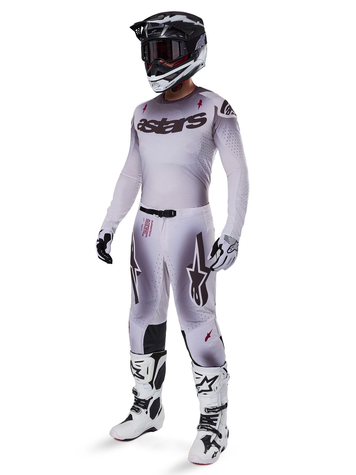 Zawodnik motocrossowy w pełnym stroju wyścigowym, szaro-biała bluza z gradientem i czarnym logo „astars”, pasujące spodnie z czerwonymi akcentami i logo gwiazdy, czarno-biały kask z ciemnymi goglami, białe buty Alpinestars Tech 10, profesjonalny strój do wyścigów off-road